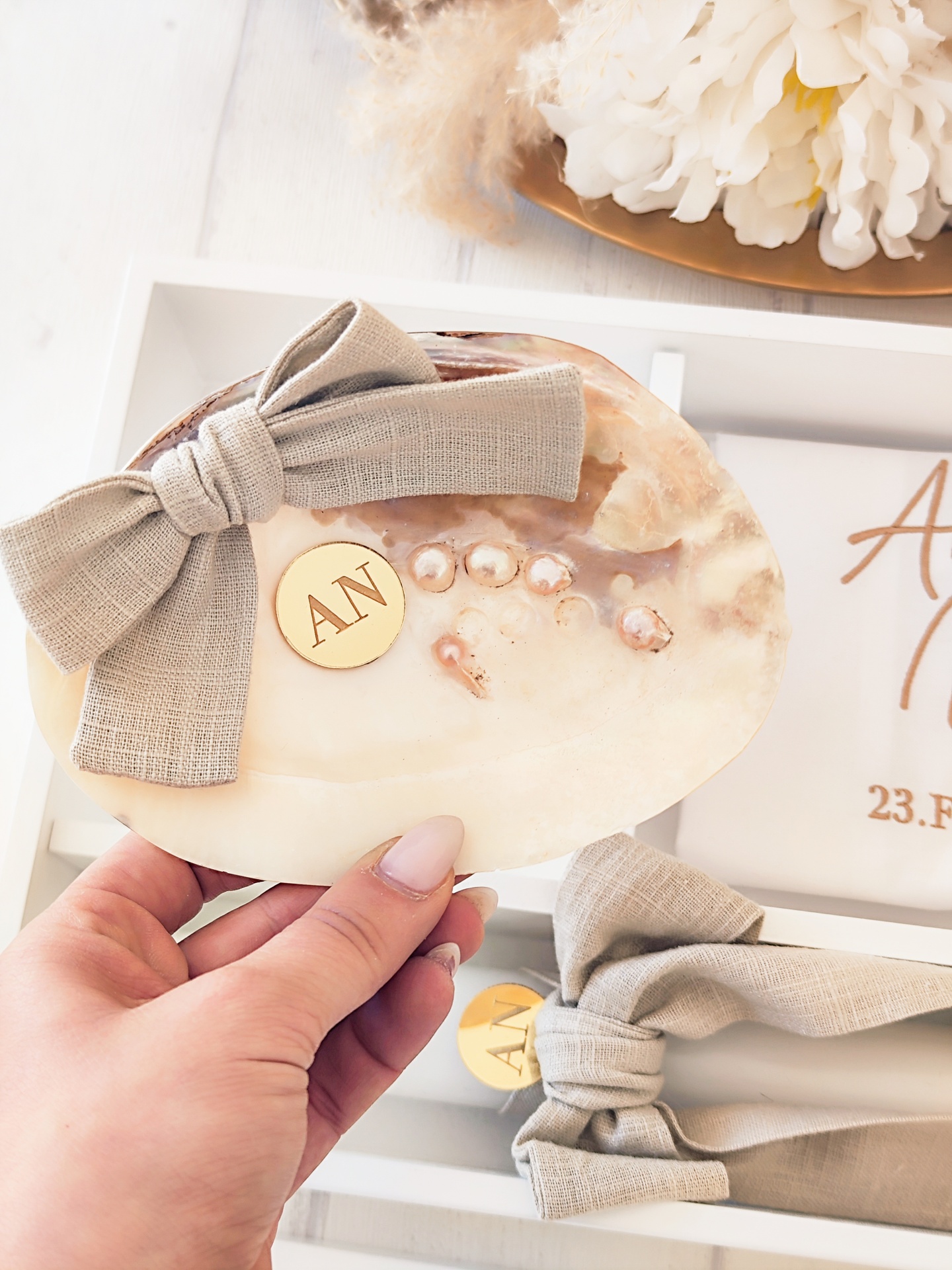 Concha decorativa com laço bege, pérolas e placa dourada com letras AN