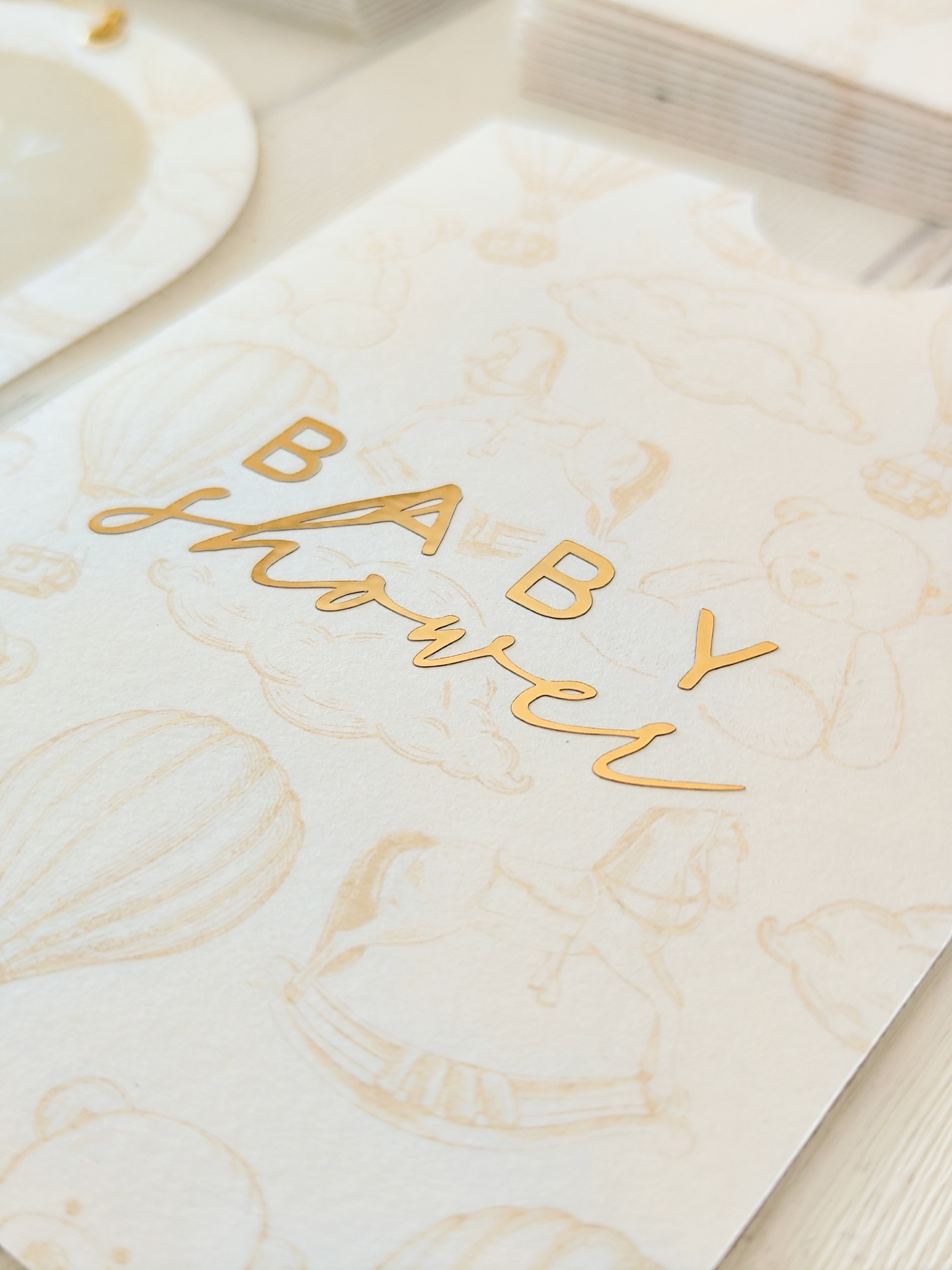 Cartão decorativo para baby shower com desenhos de balão, ursinho e cavalo de baloiço em fundo branco e letras douradas