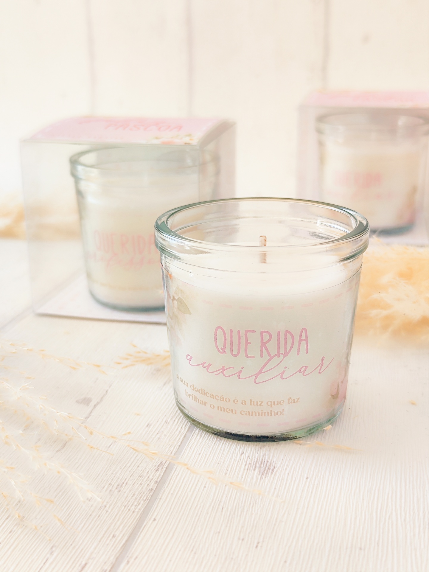 Vela em copo de vidro com texto rosa 'QUERIDA auxiliar' em cima de mesa de madeira branca
