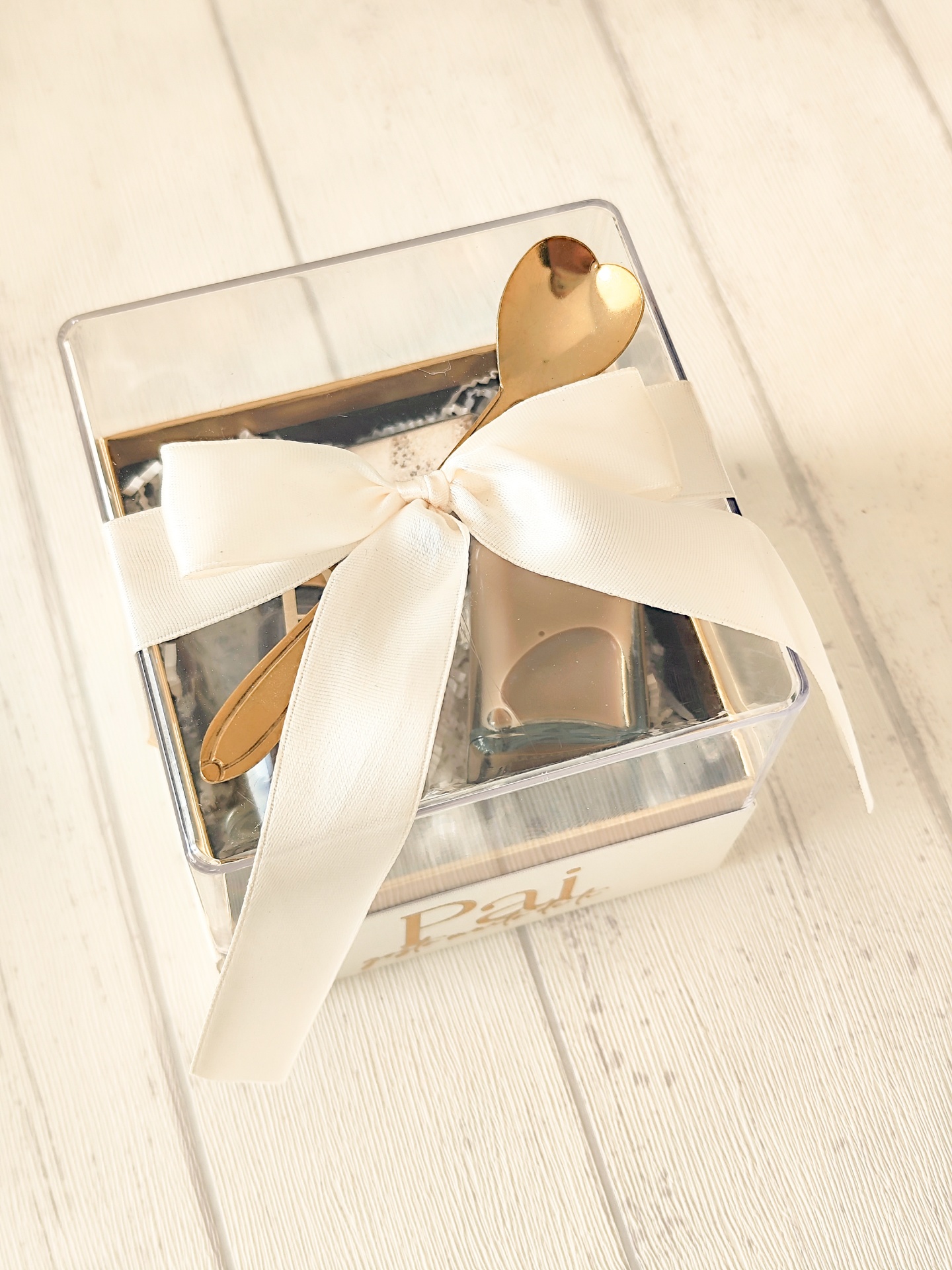Embalagem de presente transparente com laço branco e colher dourada em fundo de madeira clara