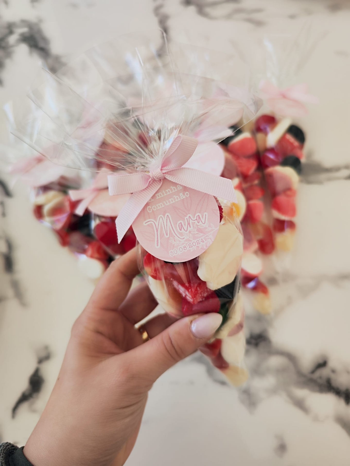 Cone transparente com doces coloridos, fita rosa e etiqueta rosa com texto, fundo marmoreado