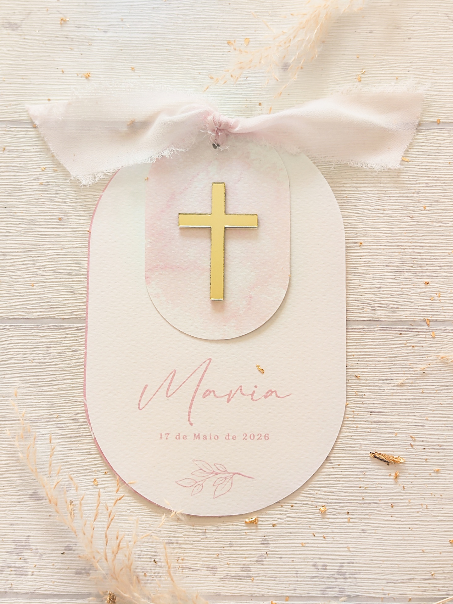 Etiqueta decorativa com cruz dourada e fita rosa claro em fundo de madeira clara