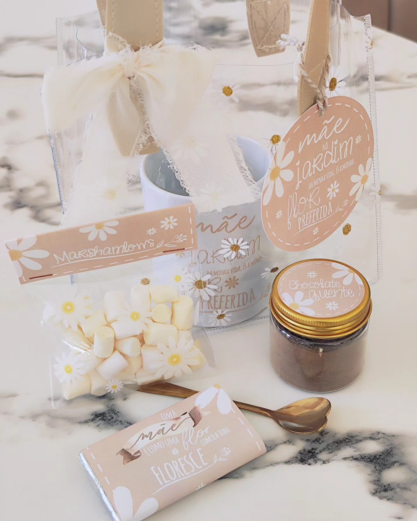 Conjunto de presente com saco transparente, marshmallows, frasco, caneca e barra de chocolate decorados com tema floral e tons rosa claro.