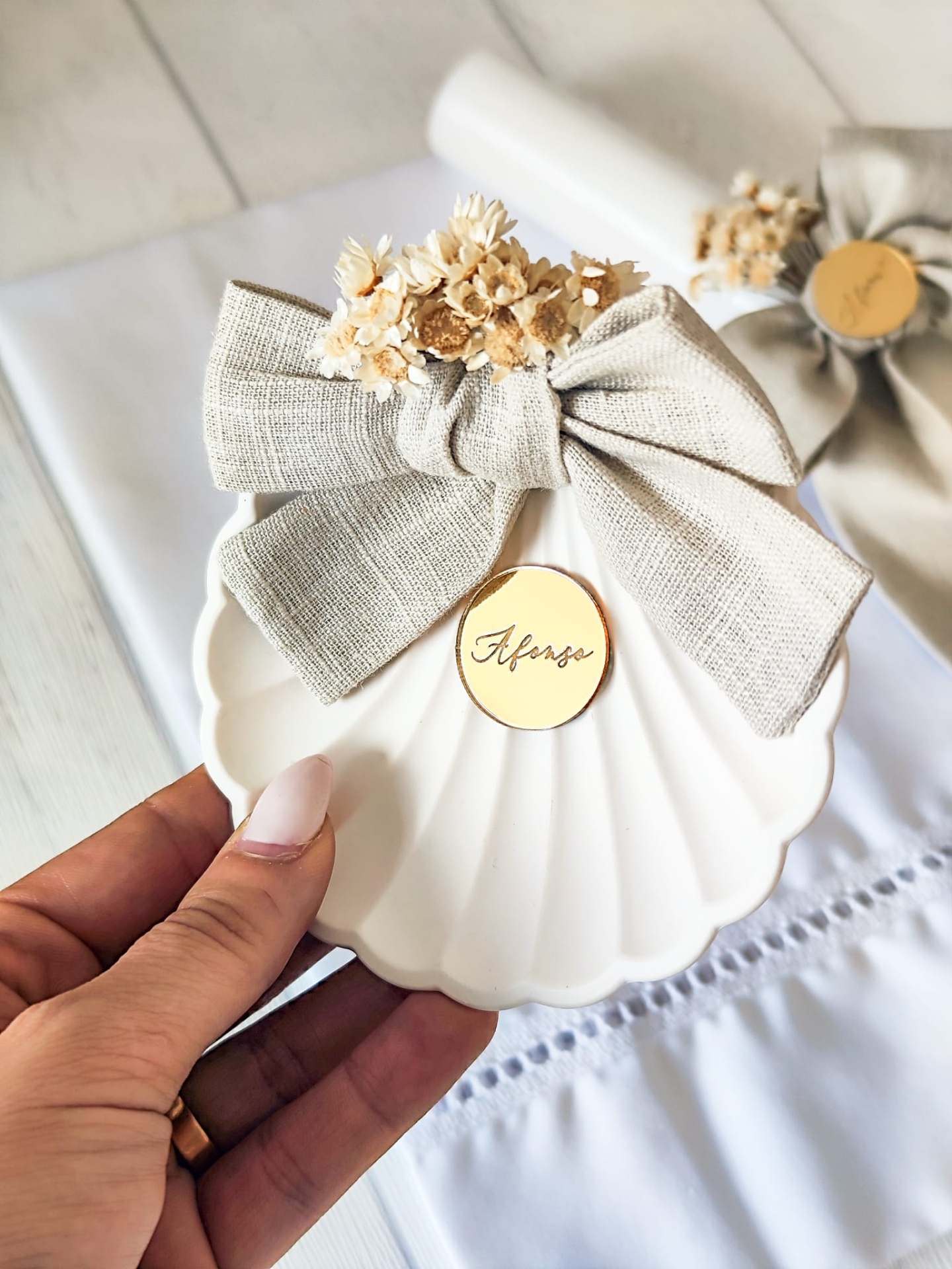 Prato decorativo branco em forma de concha com laço cinza, flores secas e etiqueta dourada 'Afonso'