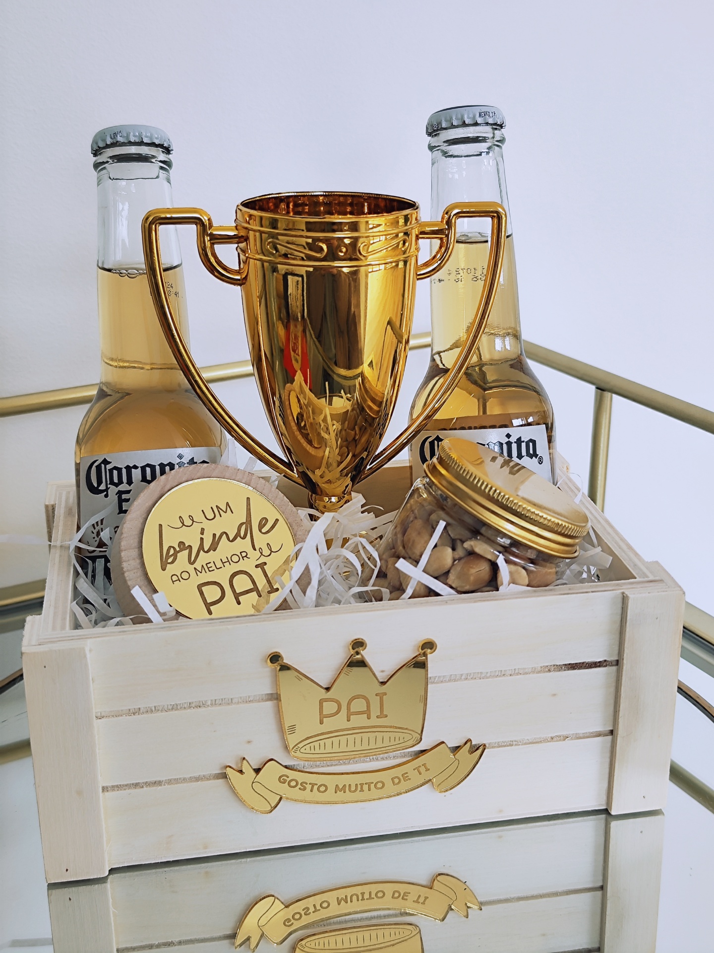 Cesta de madeira com duas garrafas de Corona, troféu dourado, frasco de amendoins e botão dourado com texto