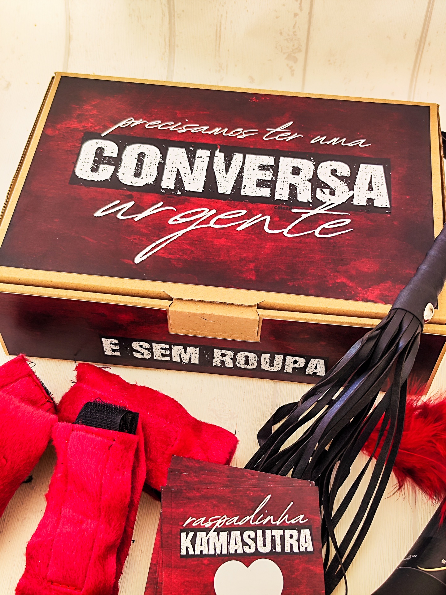 Caixa de cartão com texto, algemas vermelhas, instrumento de couro preto e livro 'raspadinha KAMASUTRA'