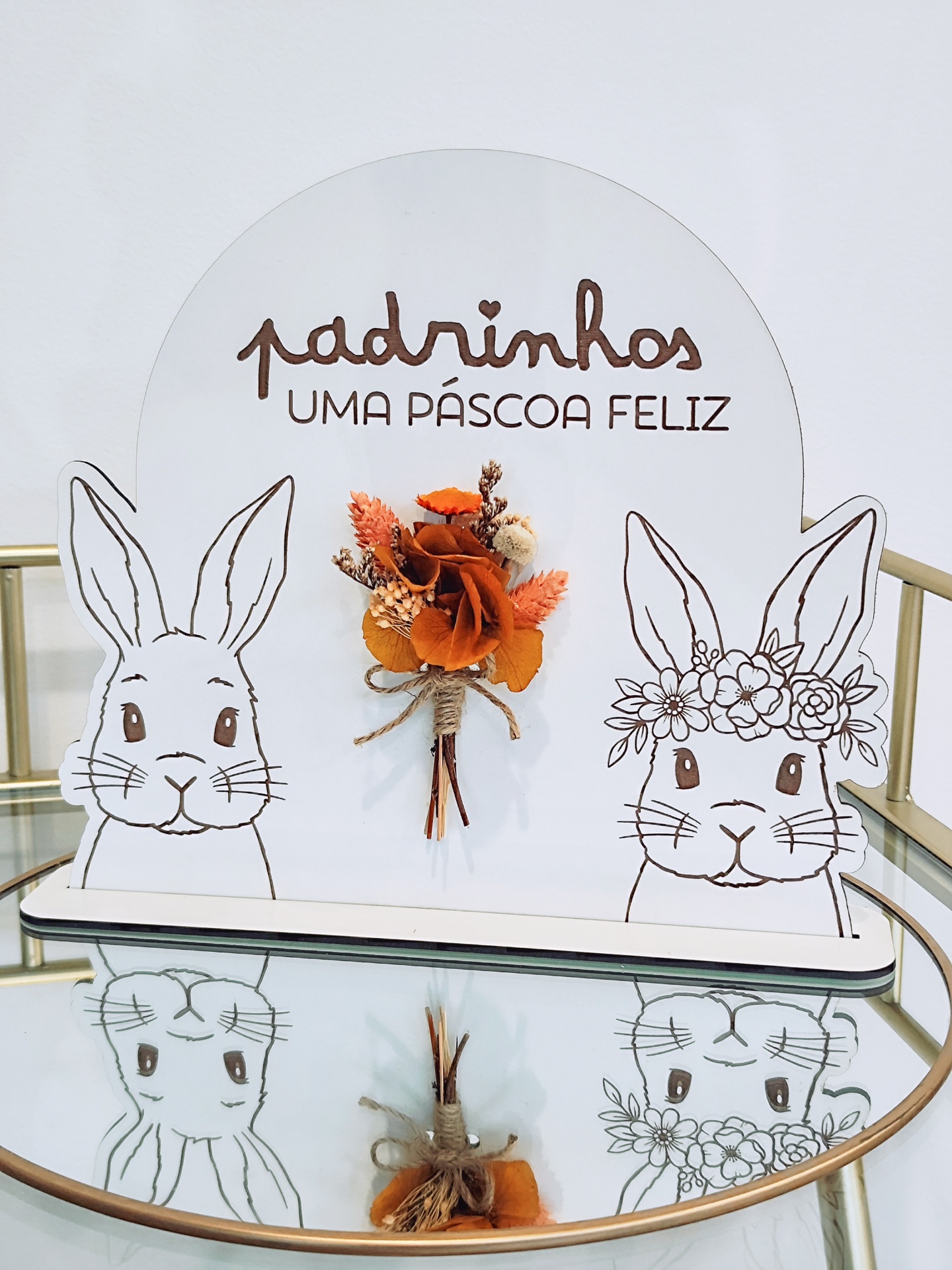 Placa decorativa branca com desenhos de coelhos e texto de Páscoa com flores secas