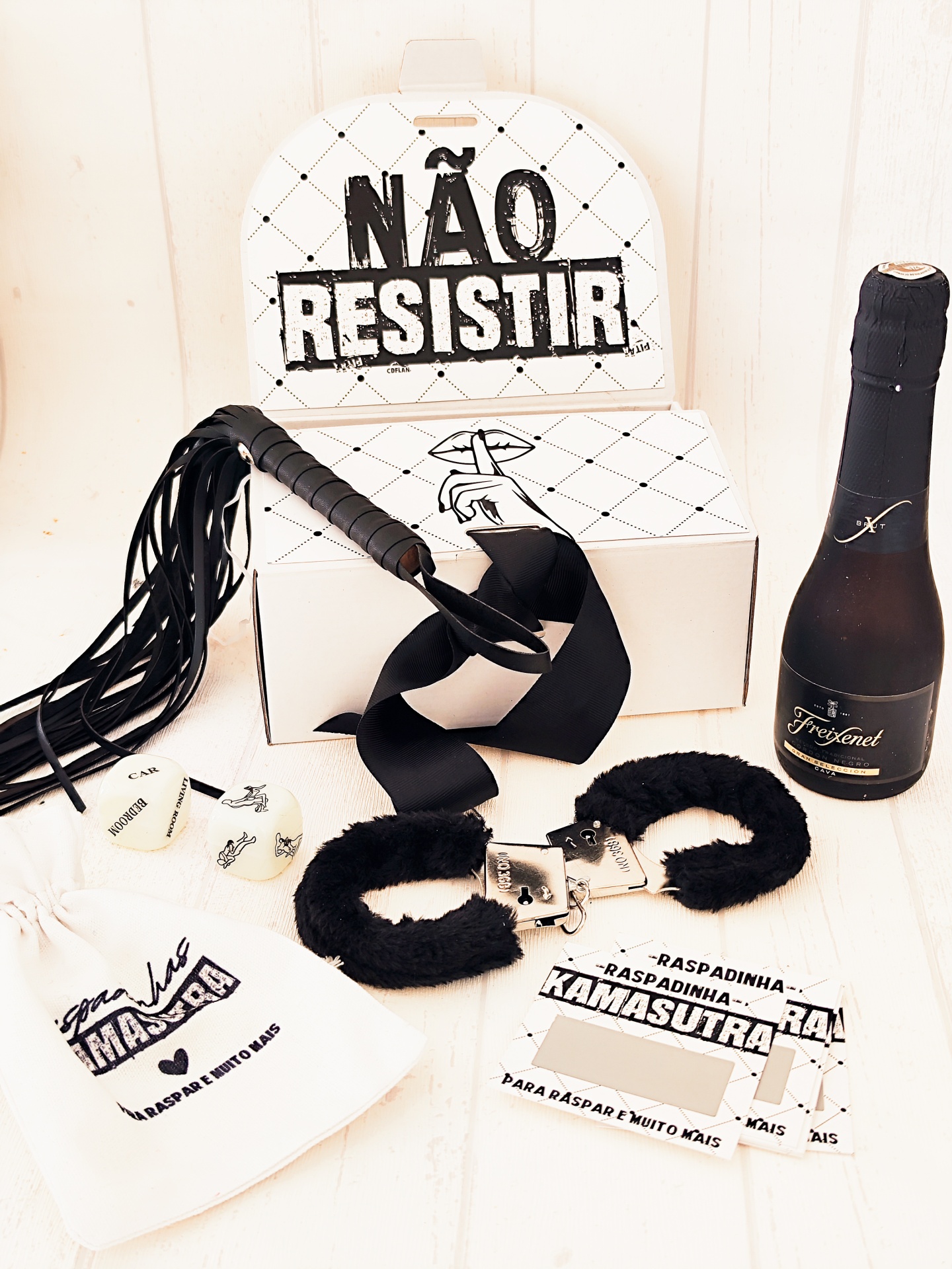 Acessórios para adultos com algemas de pelúcia, chicote e fita pretos, caixa branca com texto e garrafa de champanhe