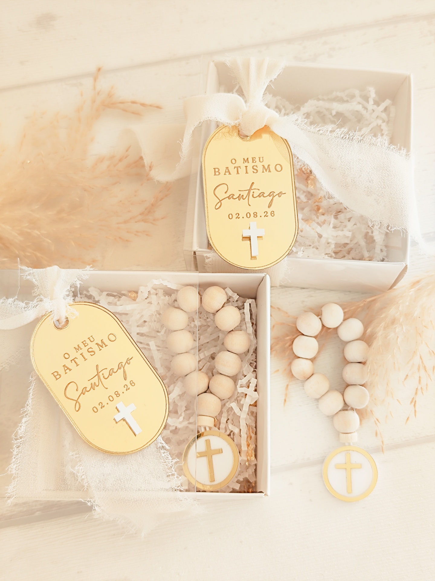 Pulseiras de contas de madeira com medalhões e etiquetas douradas para batismo, em caixas brancas com papel picado.
