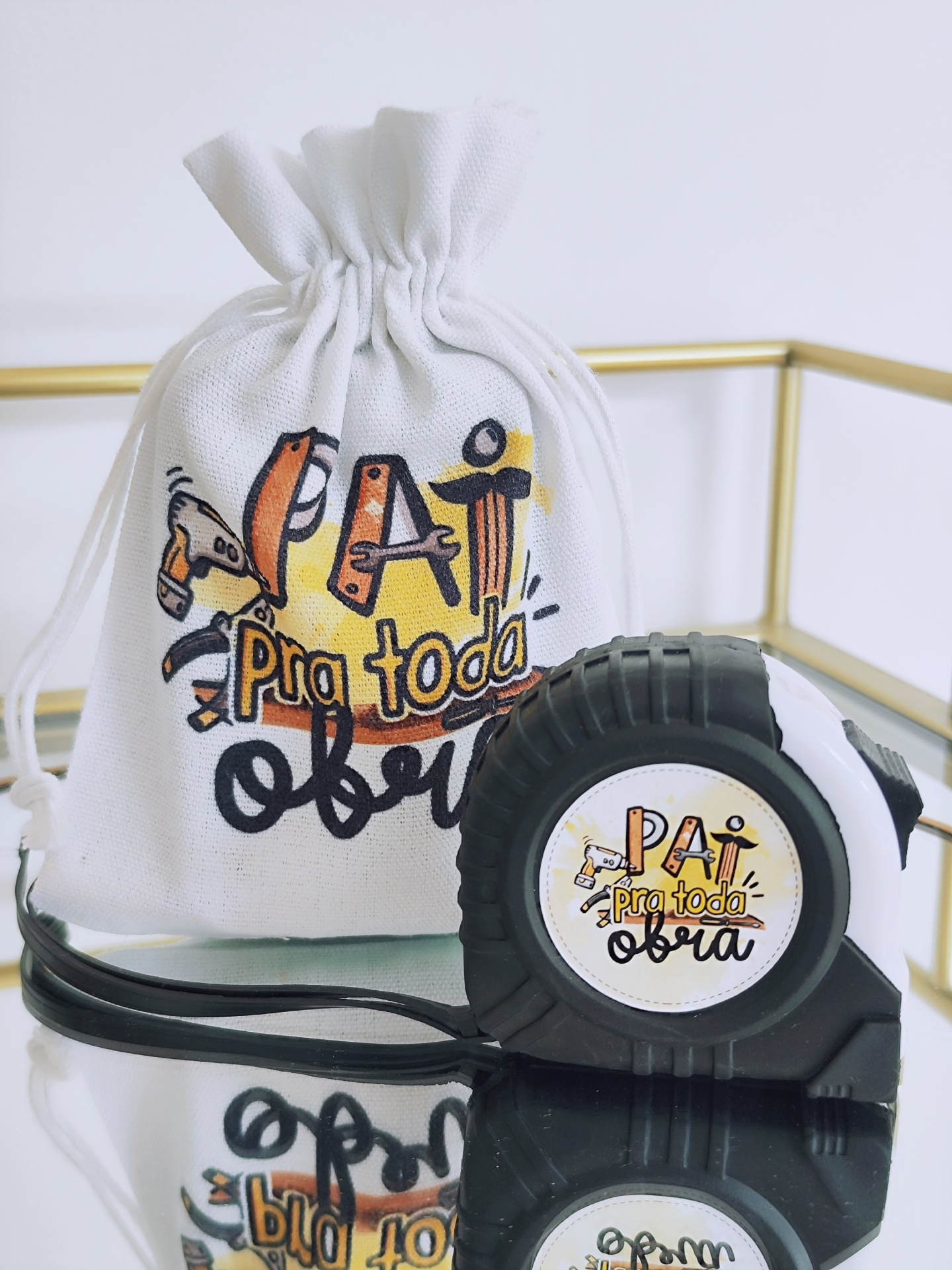 Fita métrica preta e saco branco com texto 'PAI pra toda a obra' em superfície espelhada e moldura dourada