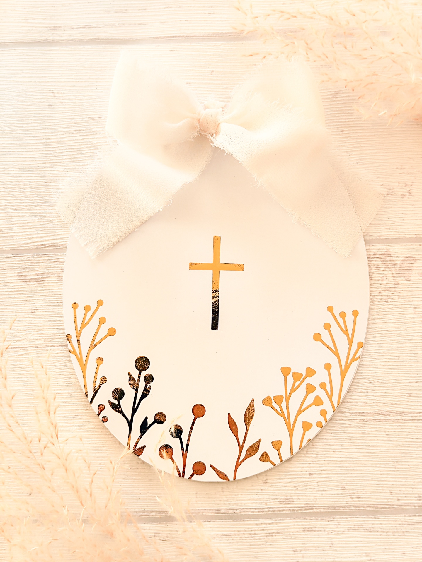 Cartão oval branco com laço e cruz dourada sobre madeira clara