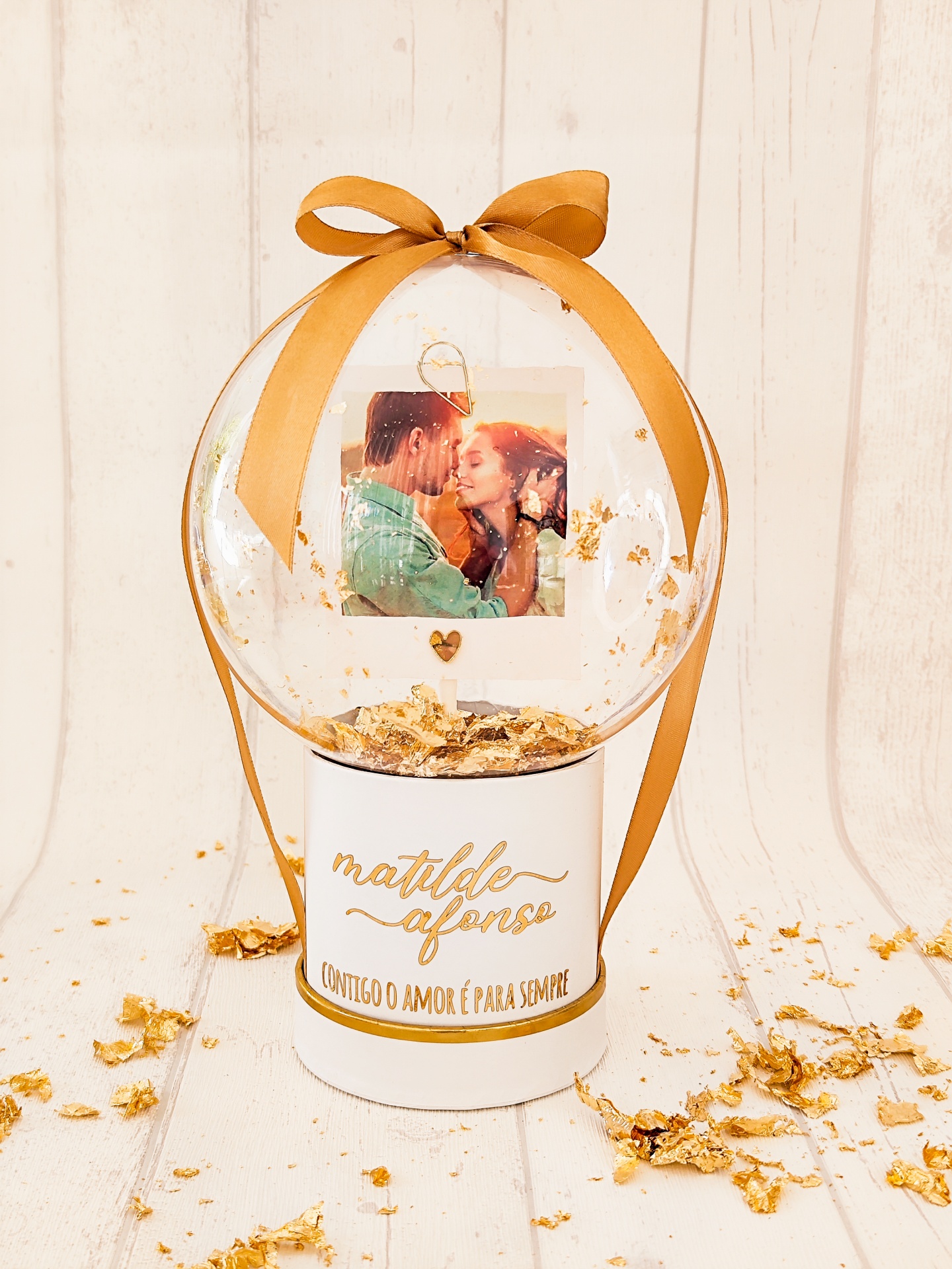 Ornamento decorativo com base branca e bola transparente contendo fotografia e folhas douradas.