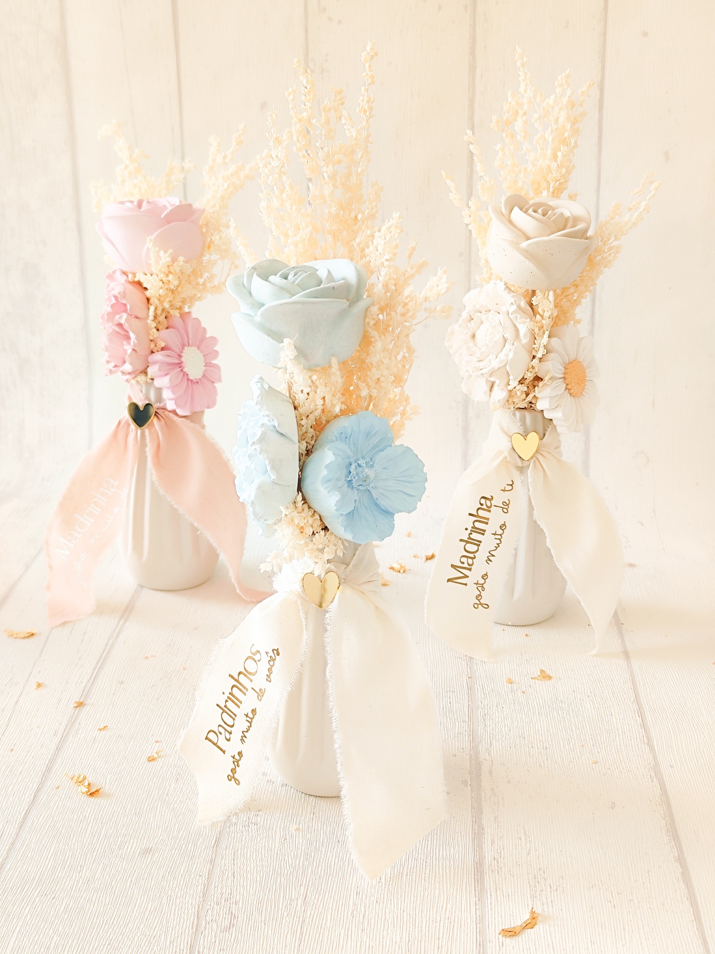 Três mini vasos decorativos com flores artificiais em tons pastel e fitas com texto em português.