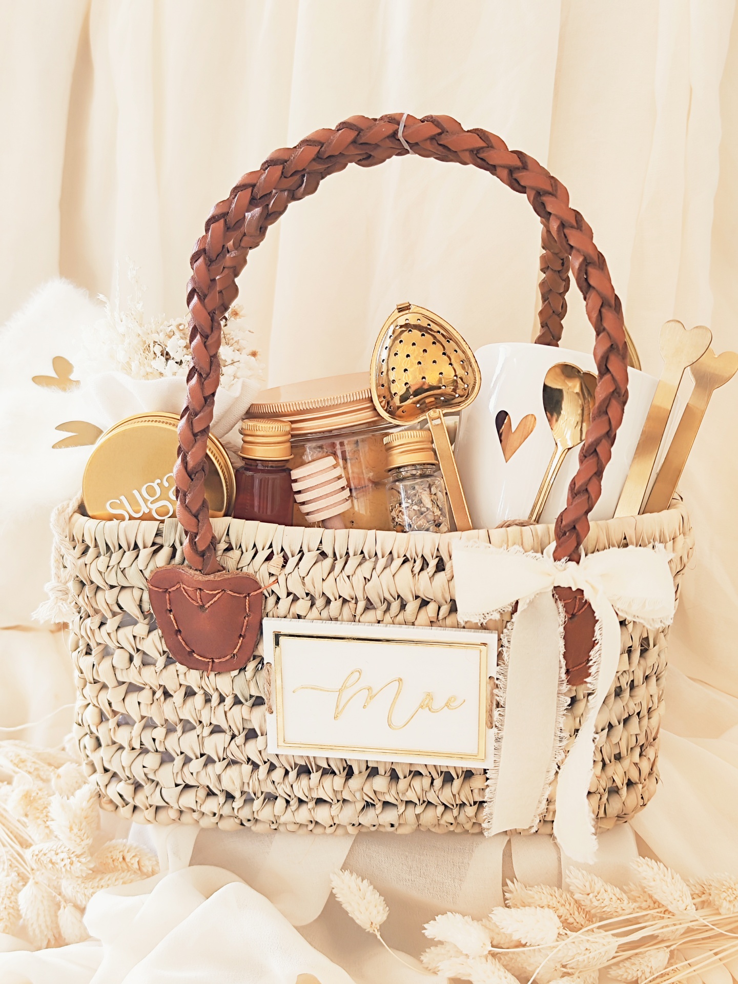 Cesto de palha com alças de couro e diversos itens para chá incluindo colher de chá em forma de coração e caneca branca com coração recortado.