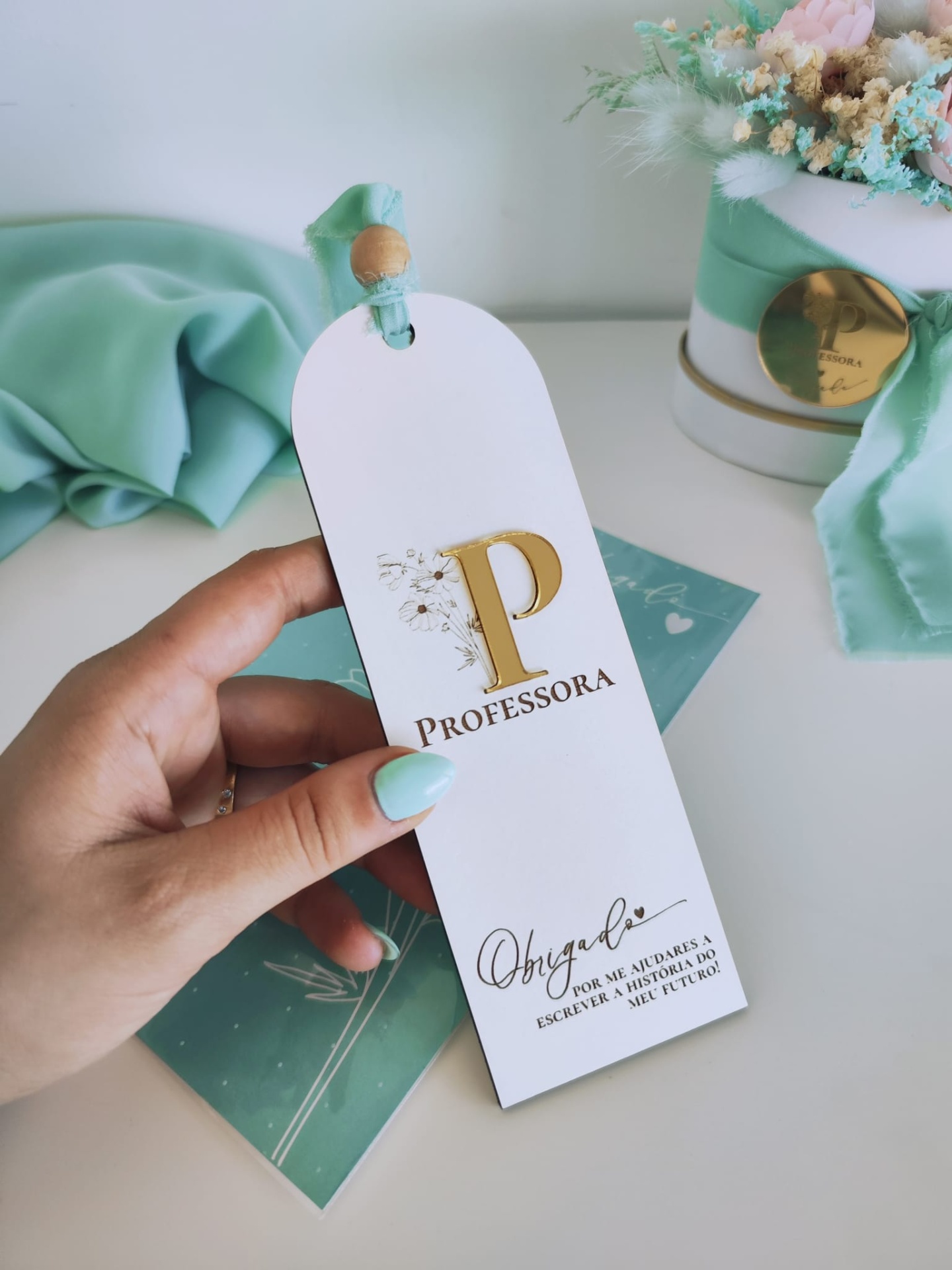 Marcador de livro branco com letra P dourada e laço verde água, mão segurando, fundo com tecido verde e caixa com flores