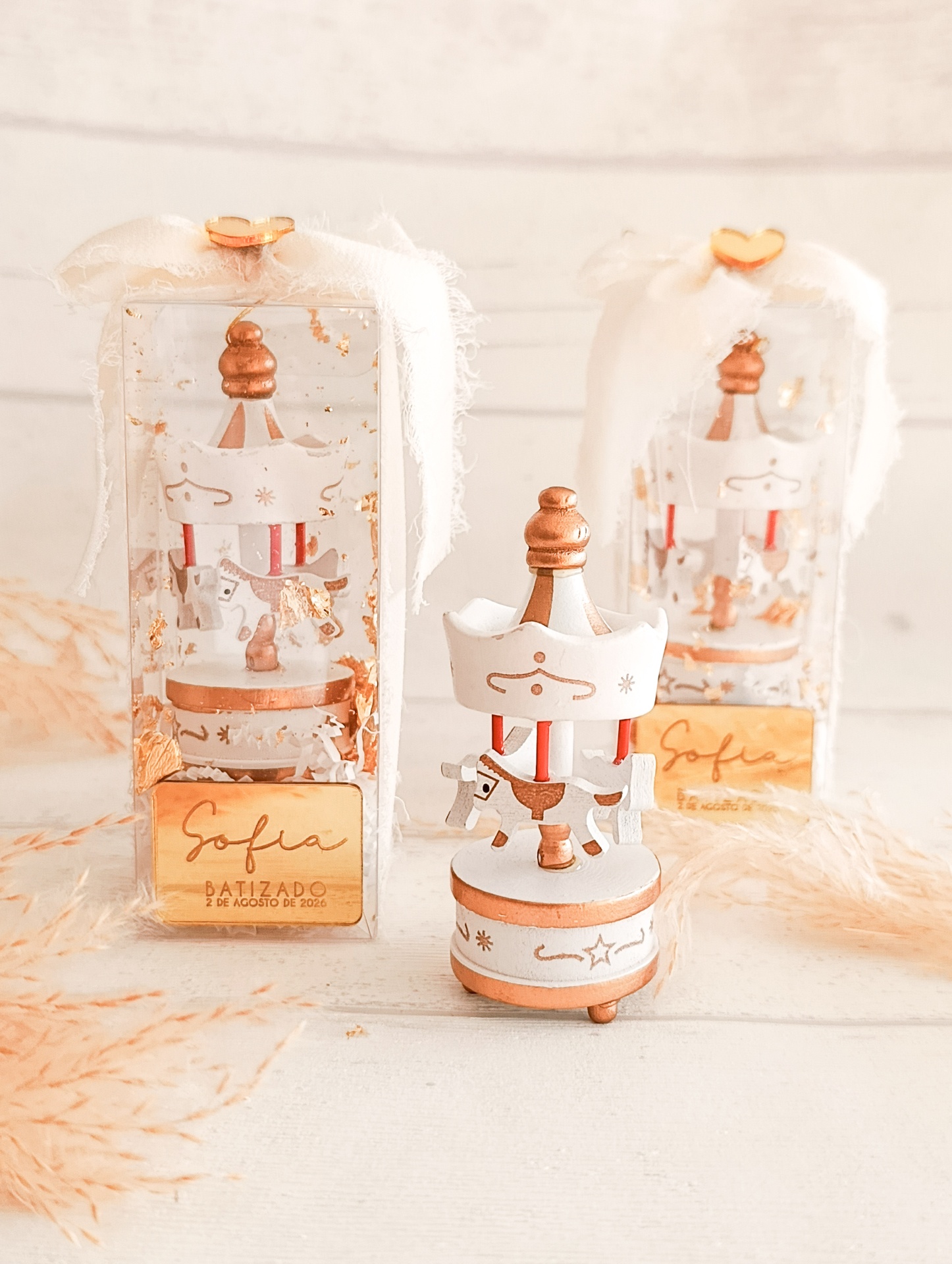 Figuras de carrossel decorativas em madeira com cavalos, laços brancos e etiquetas douradas em caixas transparentes.