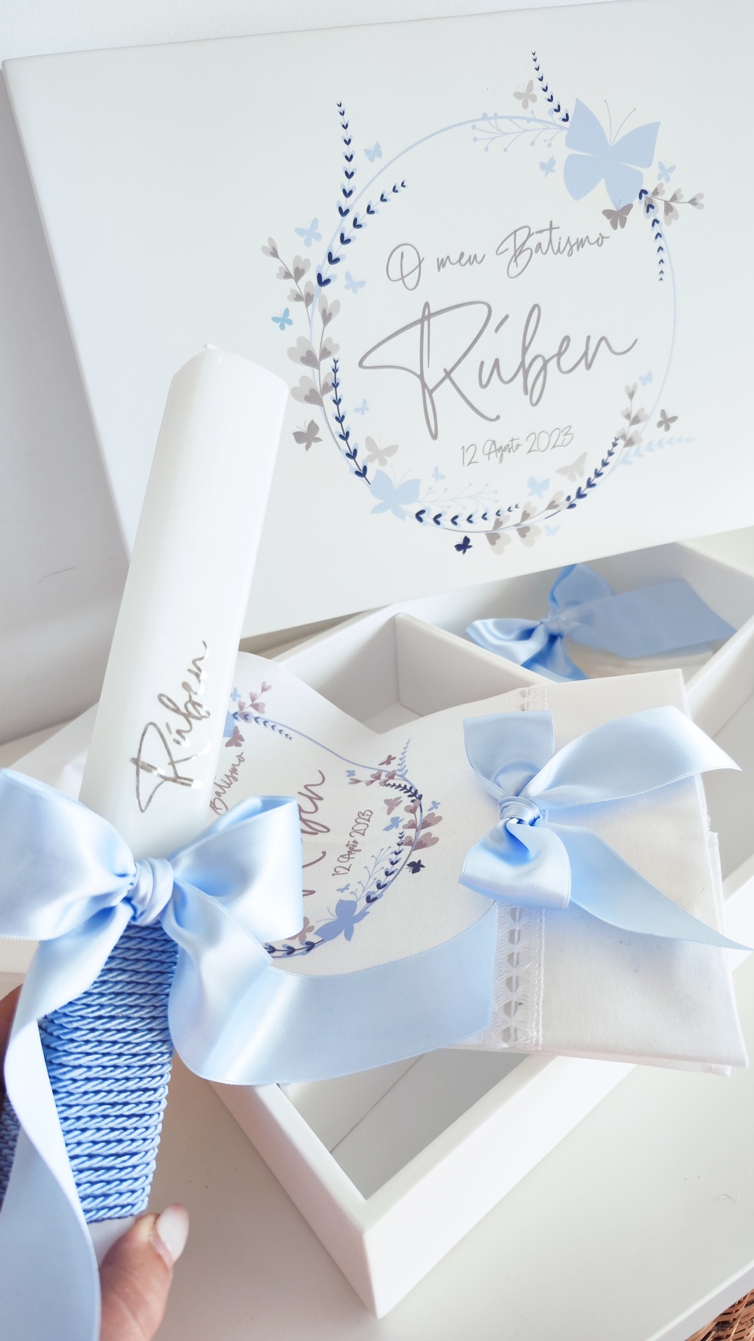 Conjunto de batismo com vela e toalha brancas e fitas azuis num estojo branco decorado