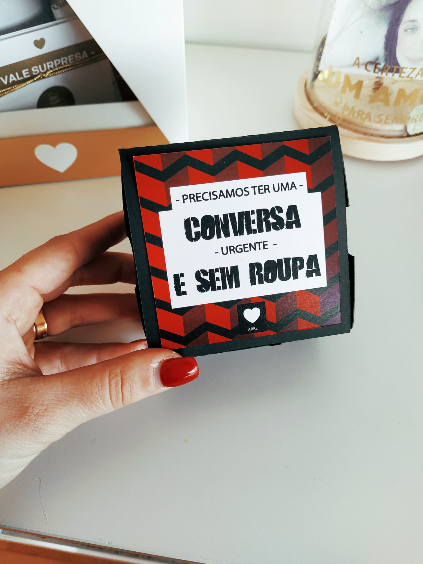 Caixa pequena vermelha e preta com texto em português segurada por mão com unha vermelha