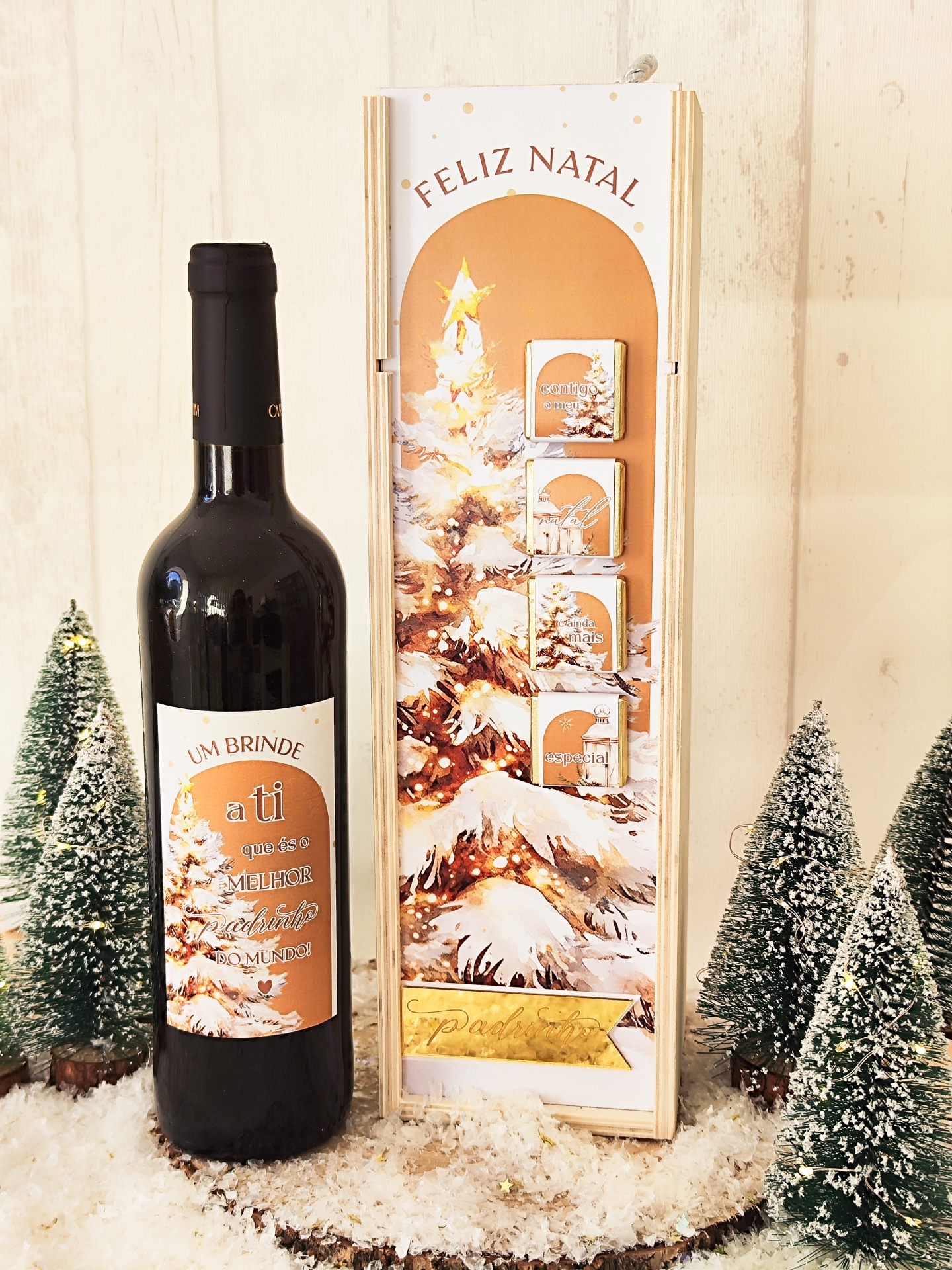 Garrafa de vinho tinto e caixa decorativa de Natal com pinheiro nevado e texto FELIZ NATAL, rodeadas por mini árvores de Natal com neve artificial.