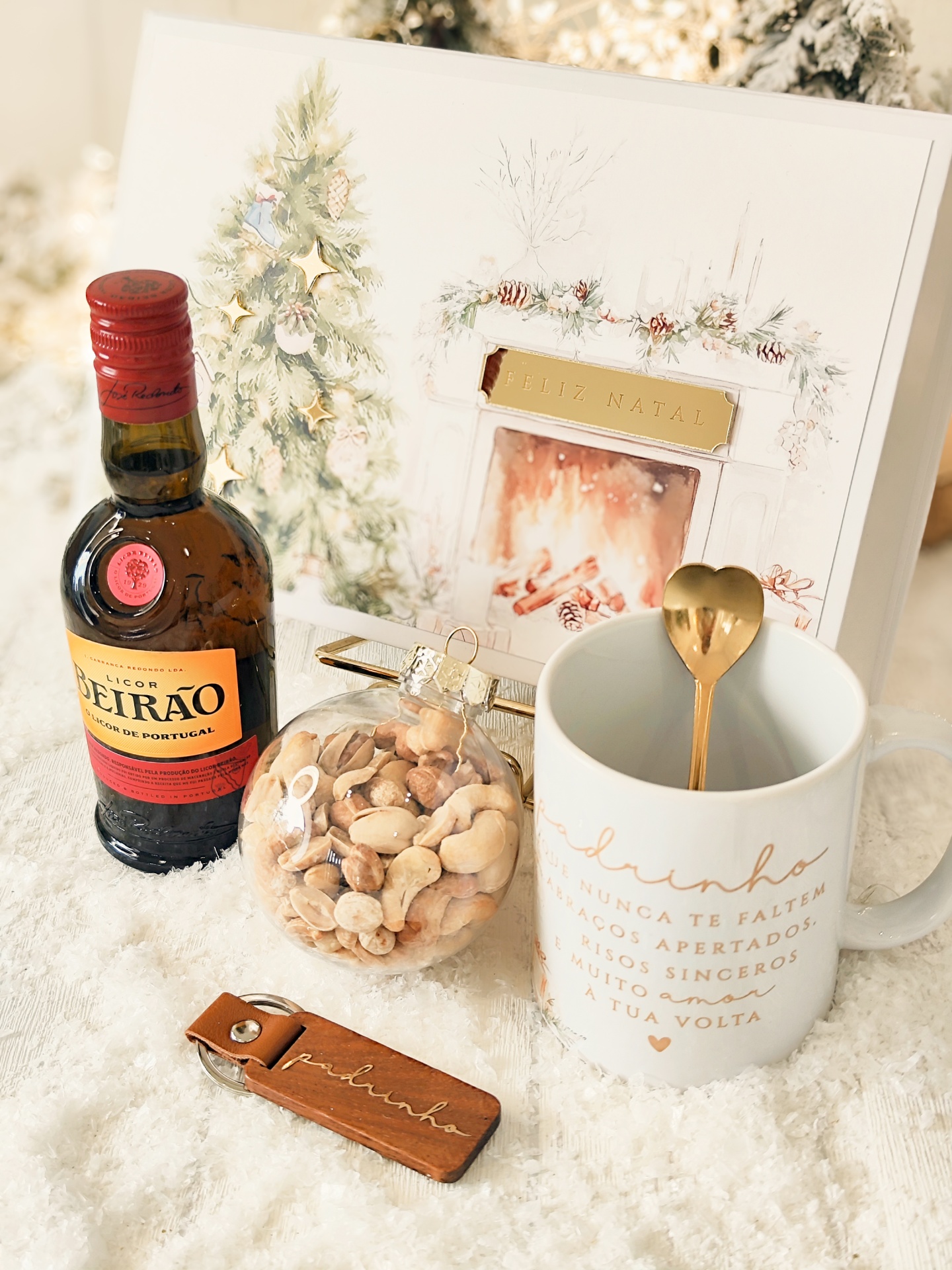 Conjunto natalício com licor, bola de frutos secos, caneca com texto e porta-chaves sobre superfície branca e cartão de Feliz Natal ao fundo