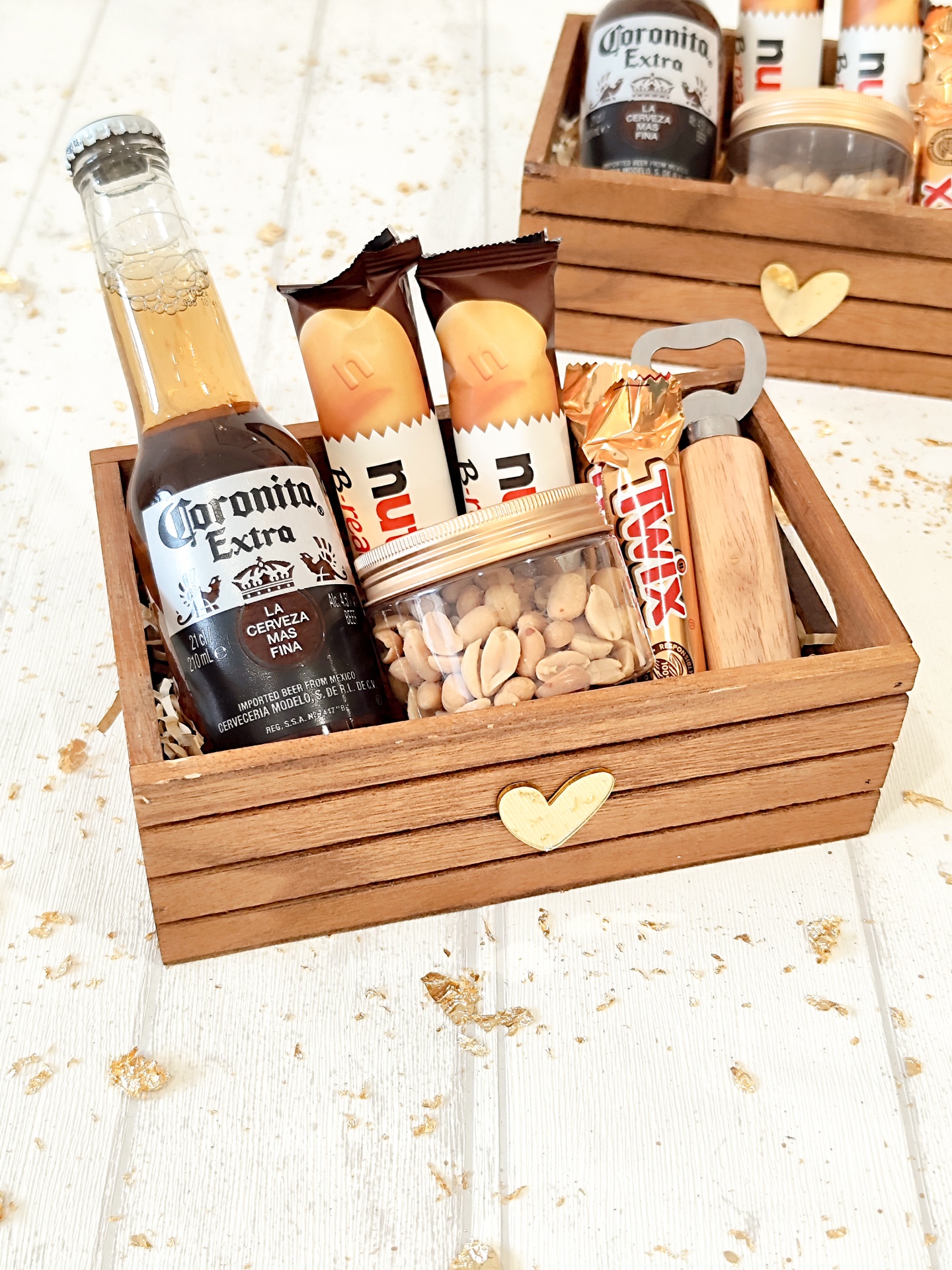 Caixa de madeira com coração dourado e produtos variados incluindo cerveja, chocolates e amendoins.