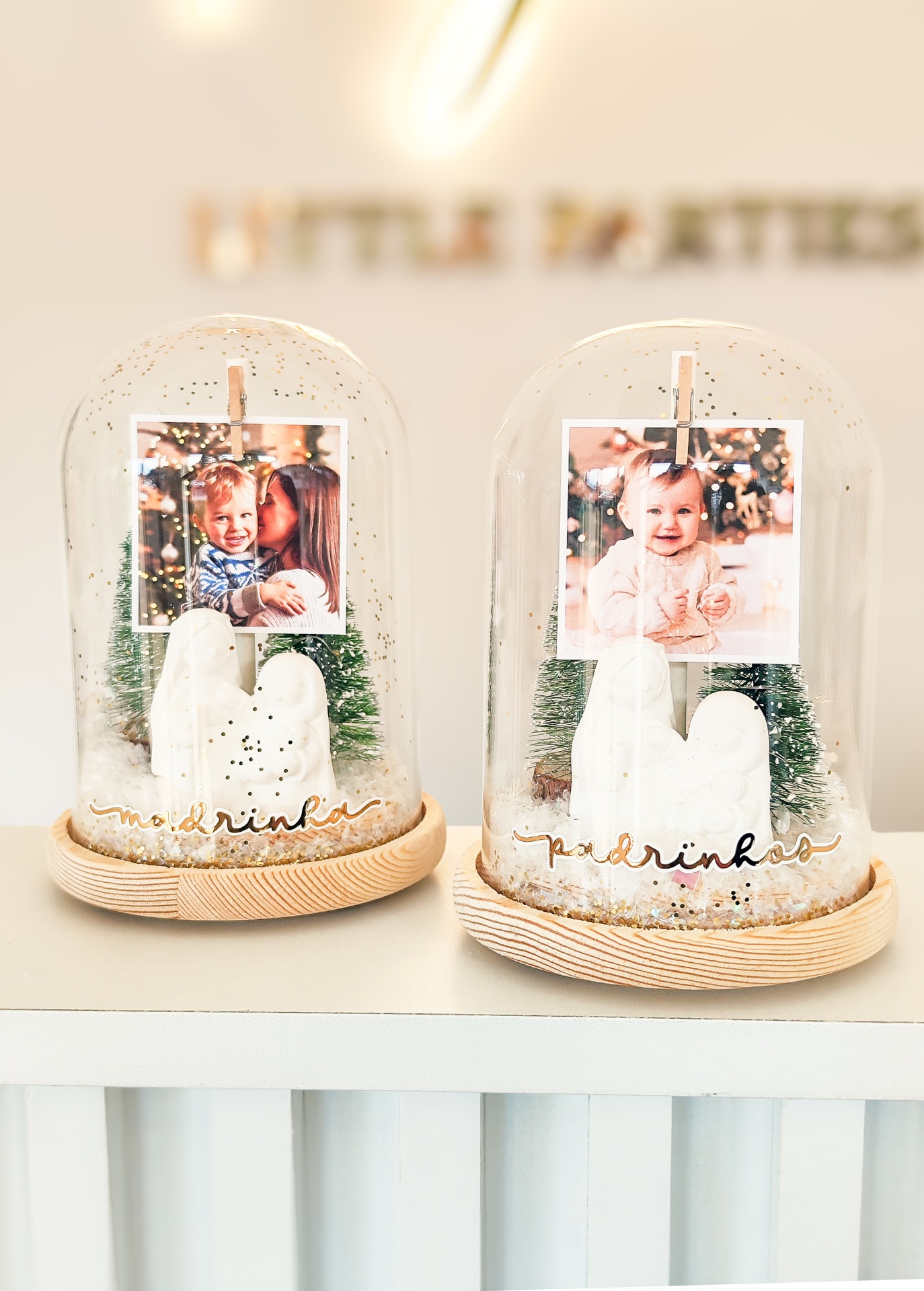 Peças decorativas em cúpulas de vidro com tema natalício, base de madeira, árvores verdes, figuras brancas e fotos de bebés.