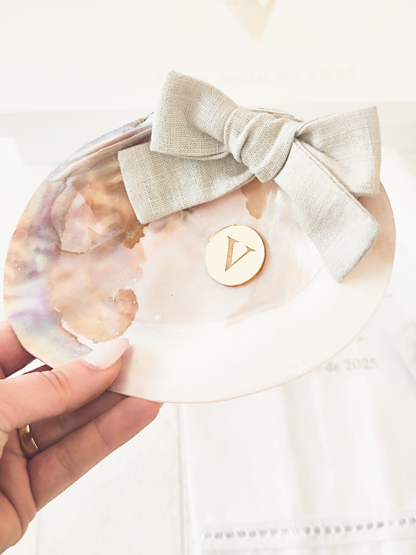 Placa decorativa em concha com laço bege e medalha dourada com letra V