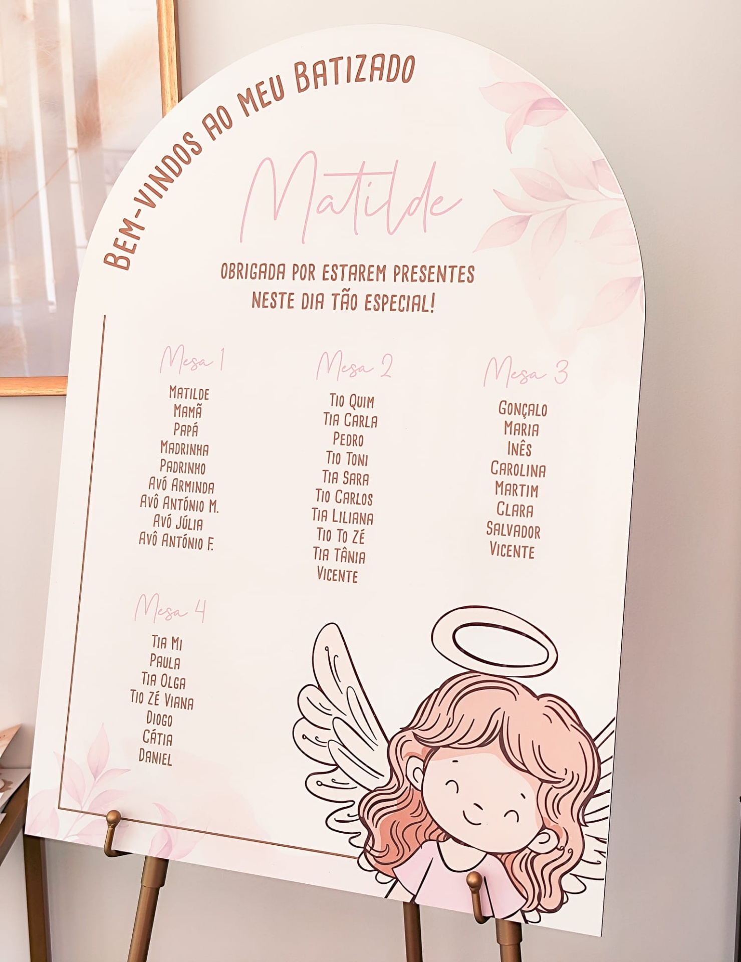 Placa branca arredondada para batizado com texto e lista de nomes, decorada com anjo desenhado, apoiada em cavalete de madeira