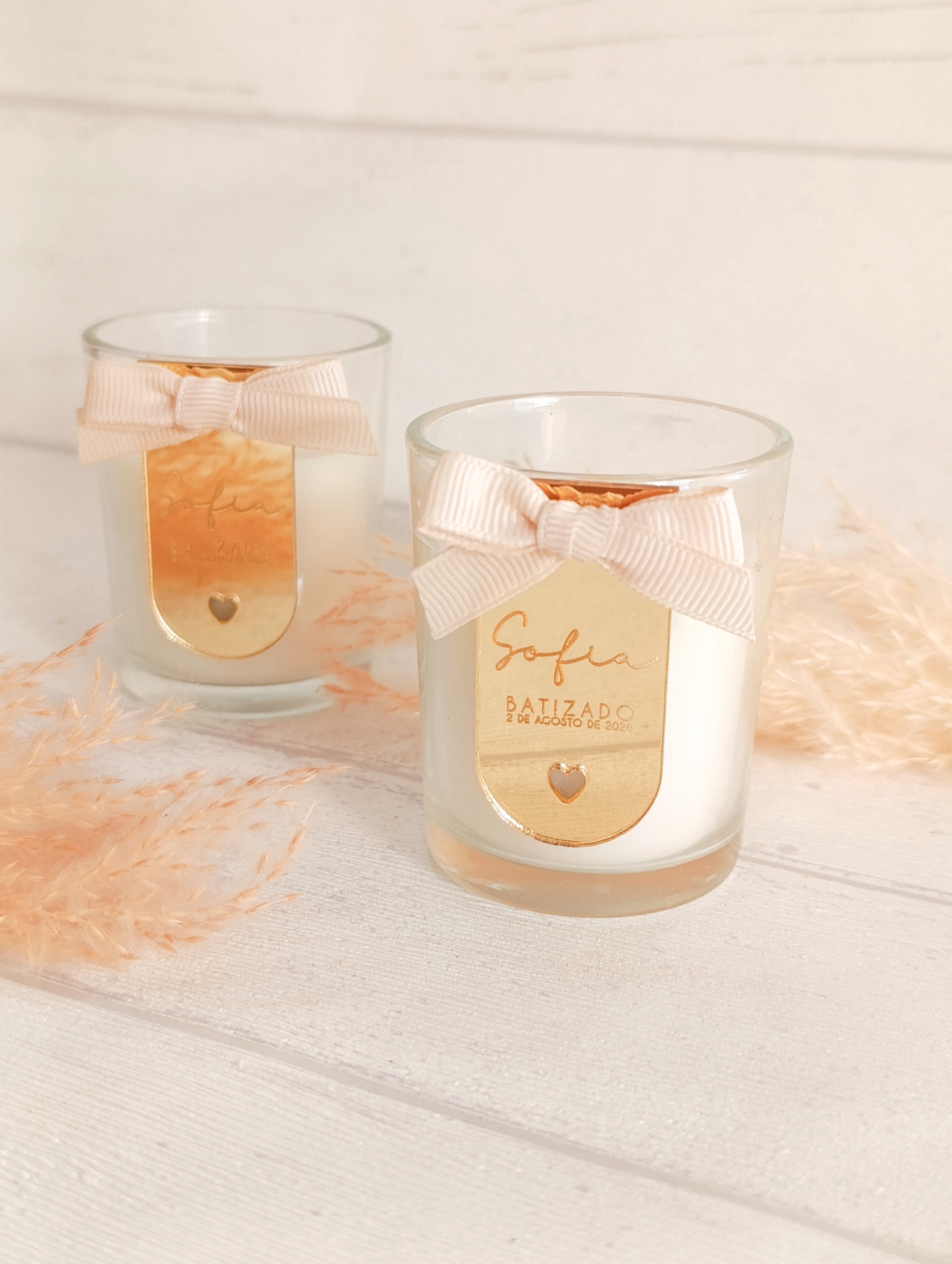 Velas em vasos de vidro com etiqueta dourada e laço rosa claro, sobre superfície branca com planta decorativa.