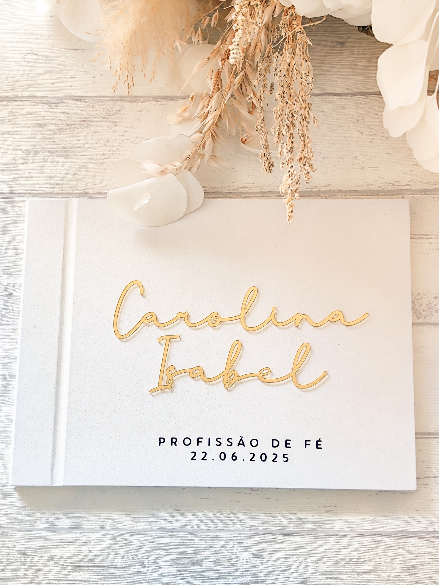 Livro branco com texto dourado e preto, decorado com flores secas sobre madeira clara