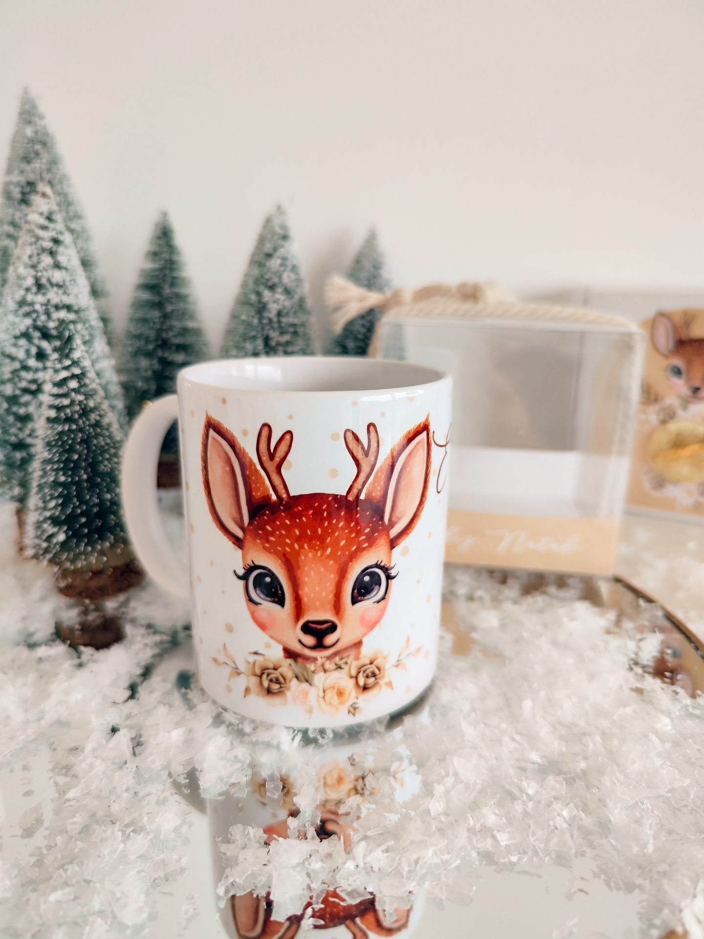 Caneca branca com desenho de veado e decoração floral, sobre superfície espelhada com neve artificial e pinheiros ao fundo.