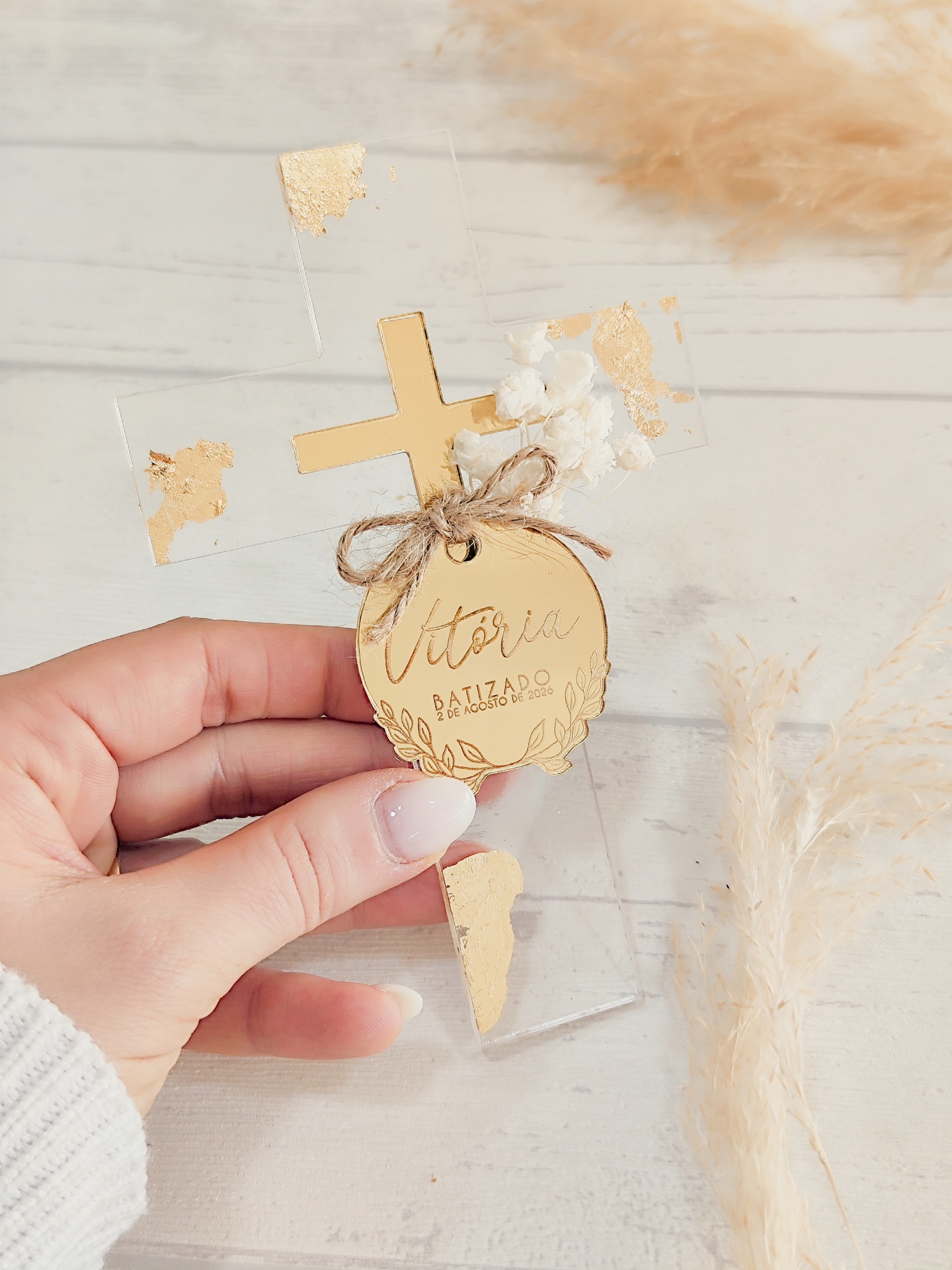Cruz decorativa transparente com detalhes dourados e etiqueta com texto para batizado.