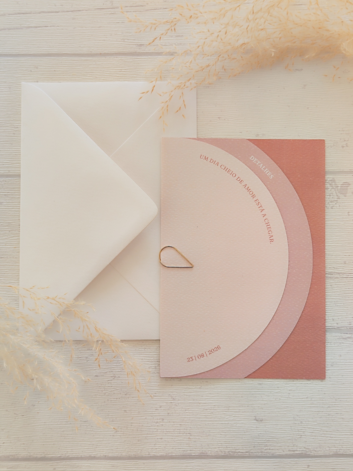 Convite rosa com envelope branco e ramos secos sobre mesa de madeira clara