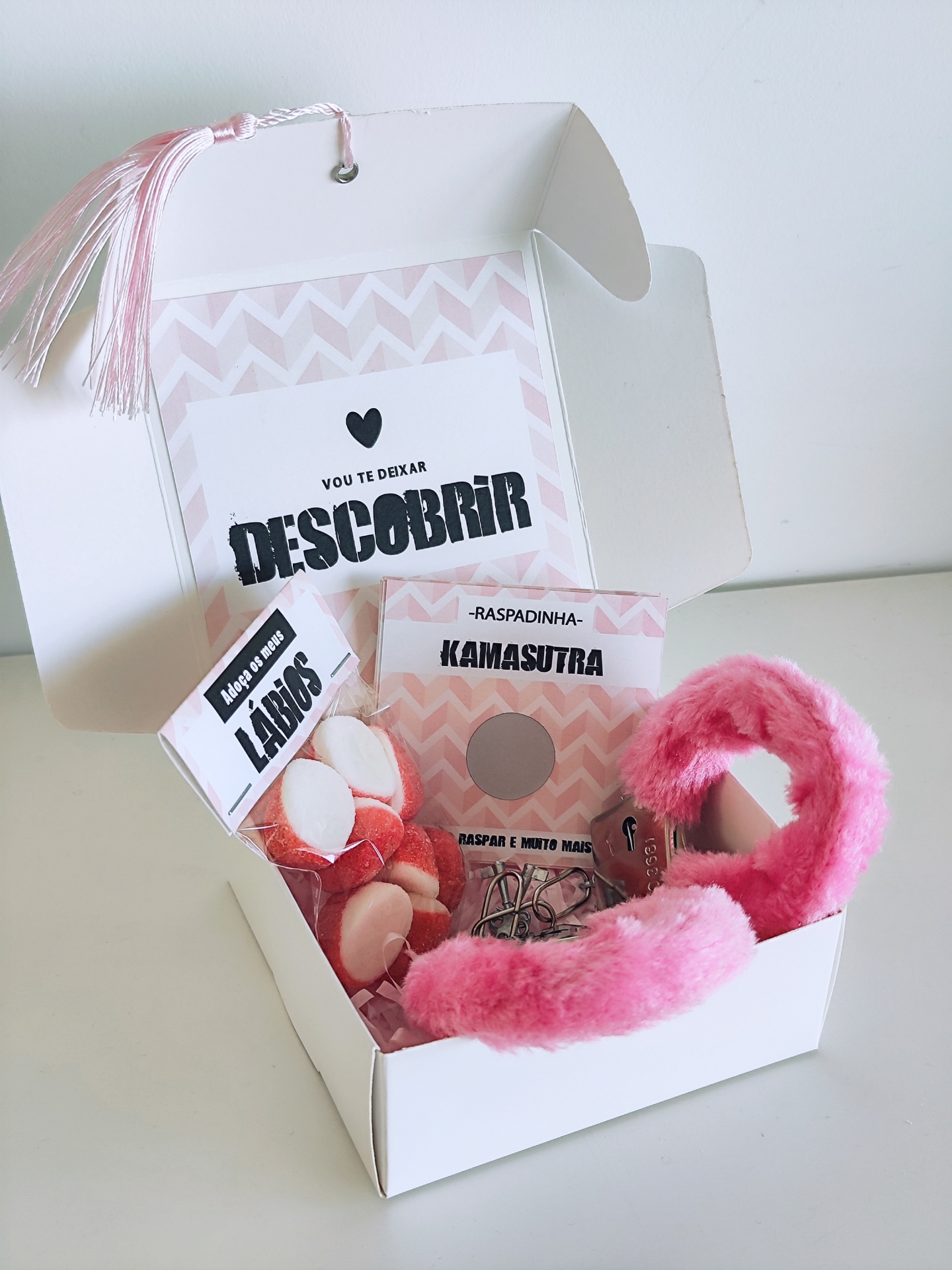 Caixa branca aberta com venda rosa felpuda, confeitos em forma de lábios e cartões com frases em português.