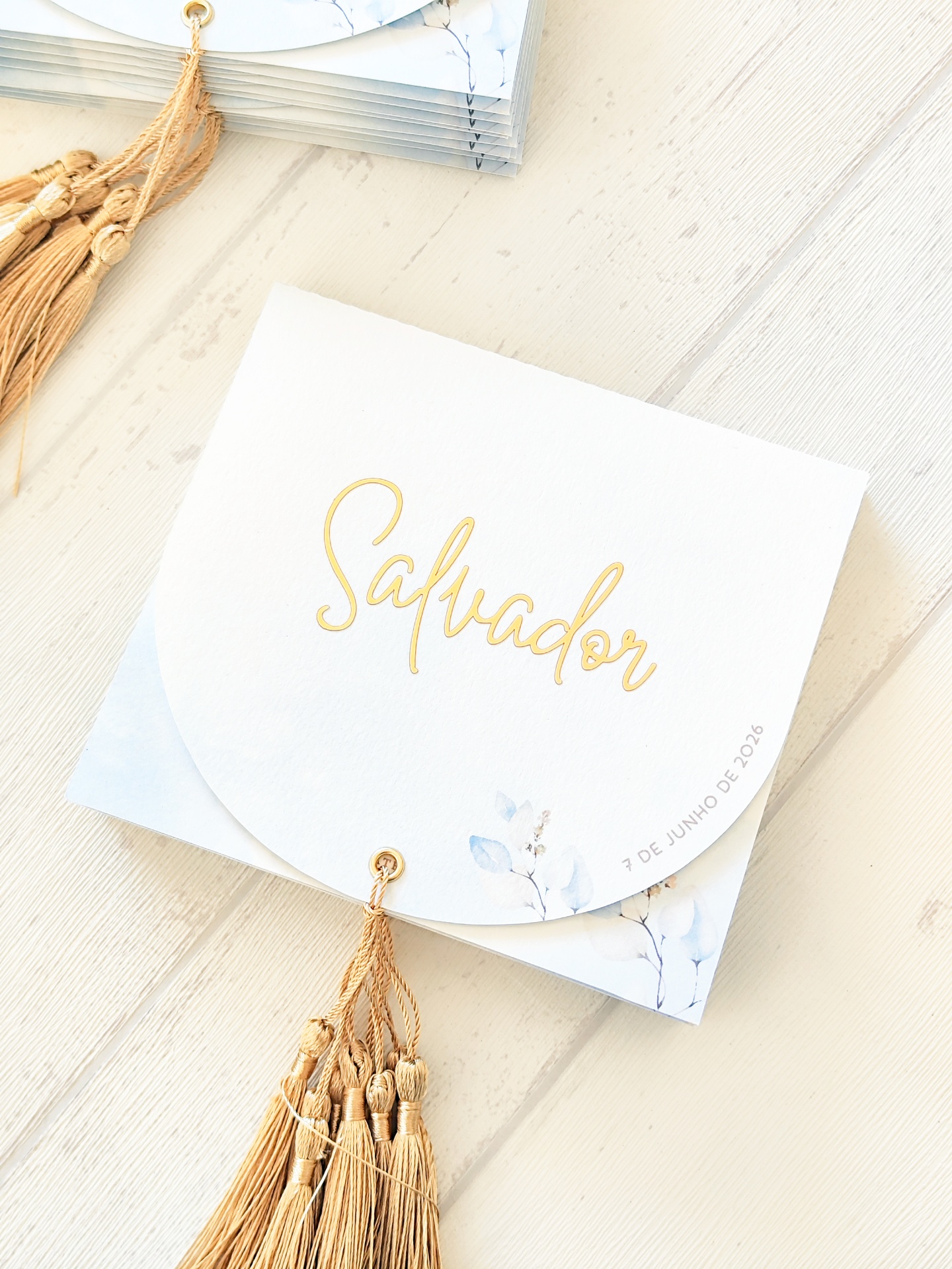 Convite branco com nome Salvador em dourado, folhas azuis e franjas douradas