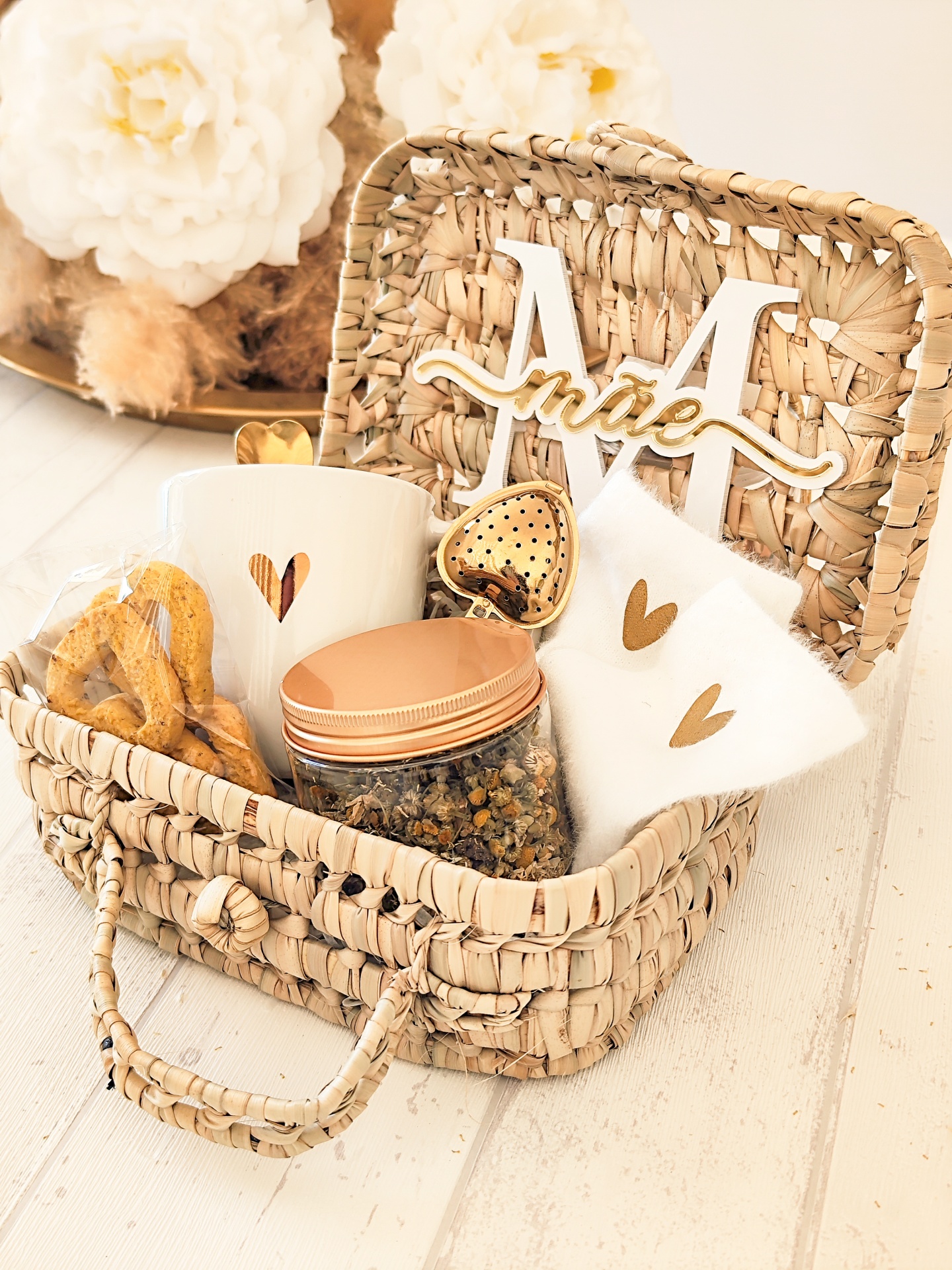 Cesto de palha com caneca branca, frasco com tampa cobre, saquinhos brancos, colher em forma de coração dourado e letra decorativa M com palavra irisé