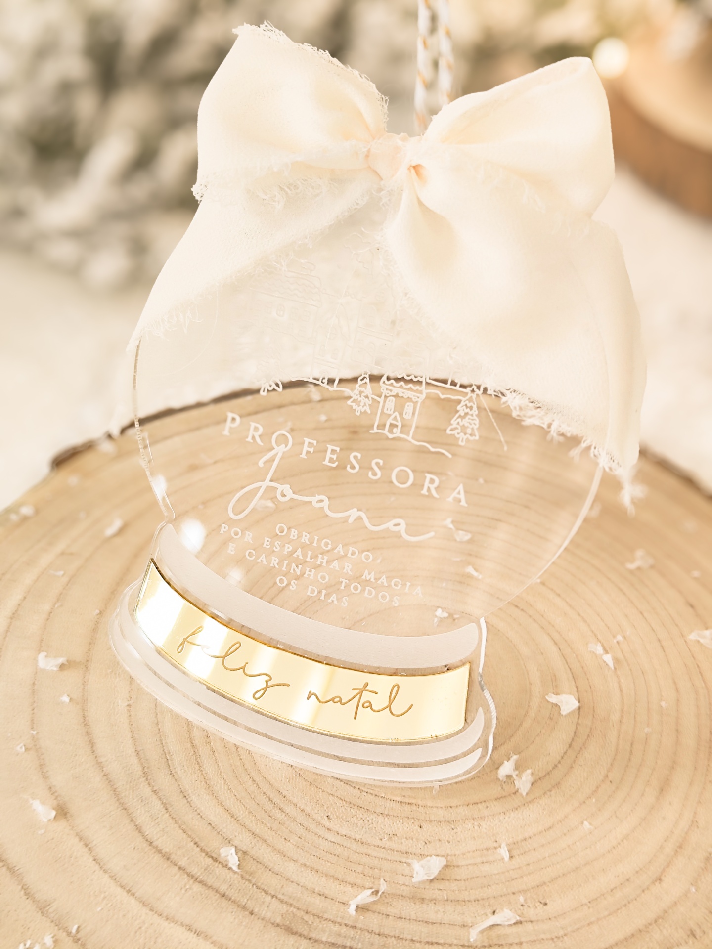 Objeto decorativo transparente redondo com laço creme e texto gravado para professora, sobre tronco de árvore