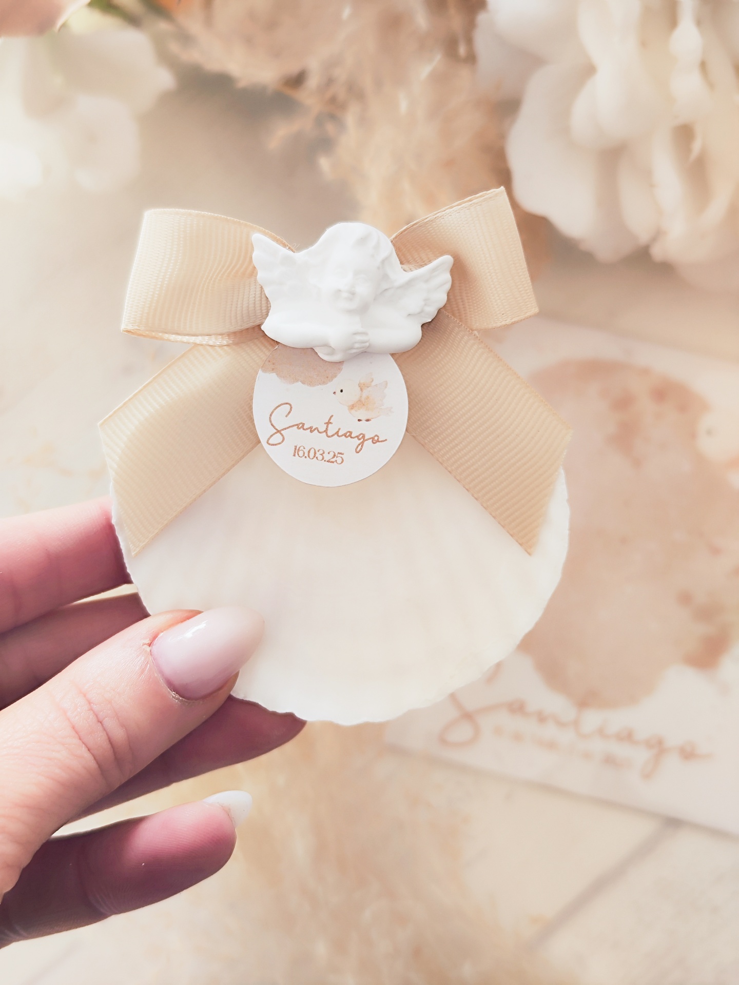 Concha marinha decorada com laço bege, anjo branco e etiqueta 'Santiago 16 03 25'