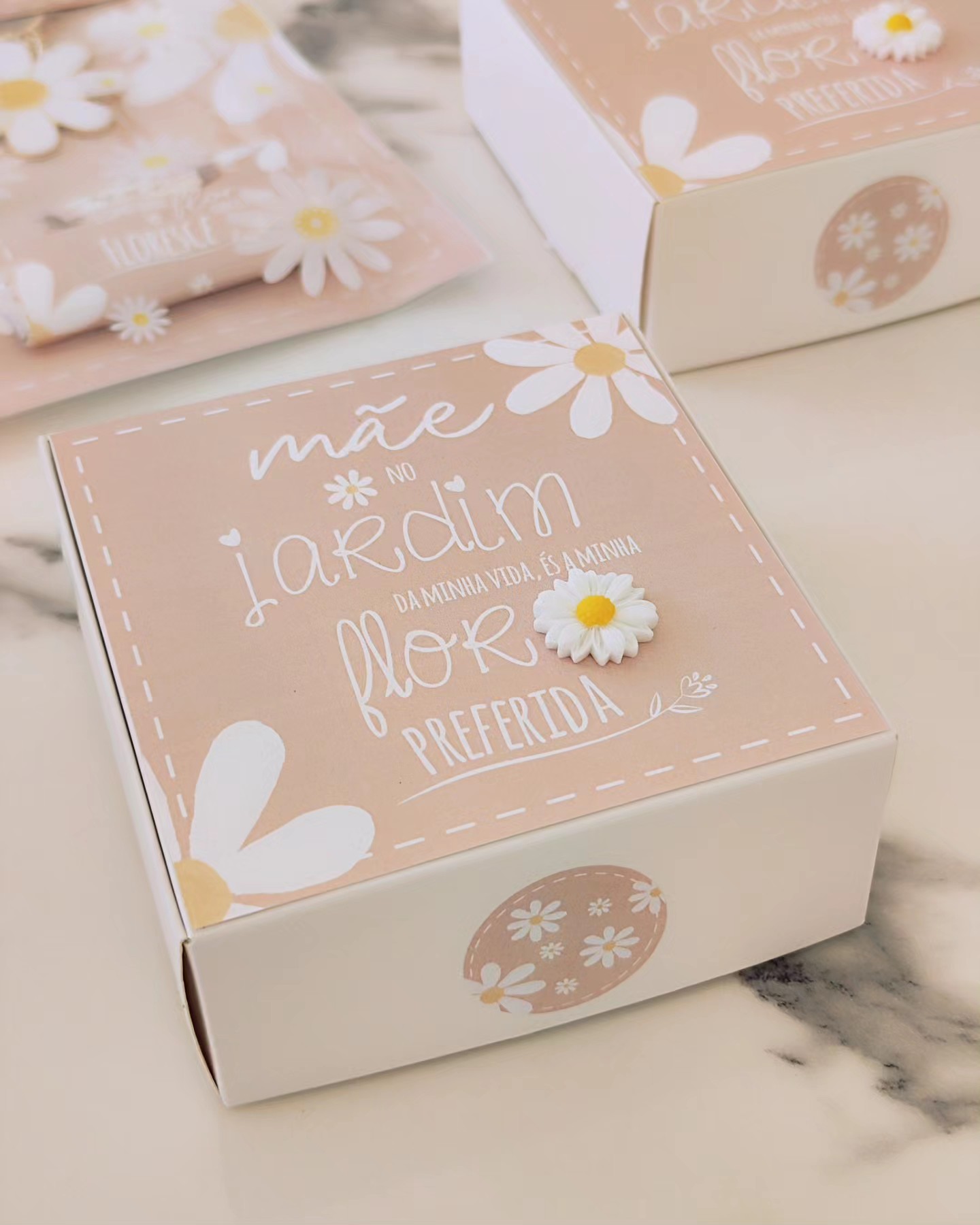 Caixa decorativa rosa claro com texto e flores brancas, incluindo uma margarida em relevo, sobre superfície marmoreada clara.