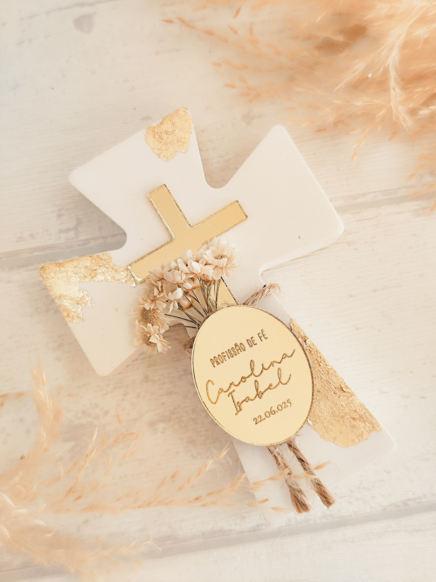 Cruz branca e dourada com flores secas e etiqueta personalizada sobre mesa clara