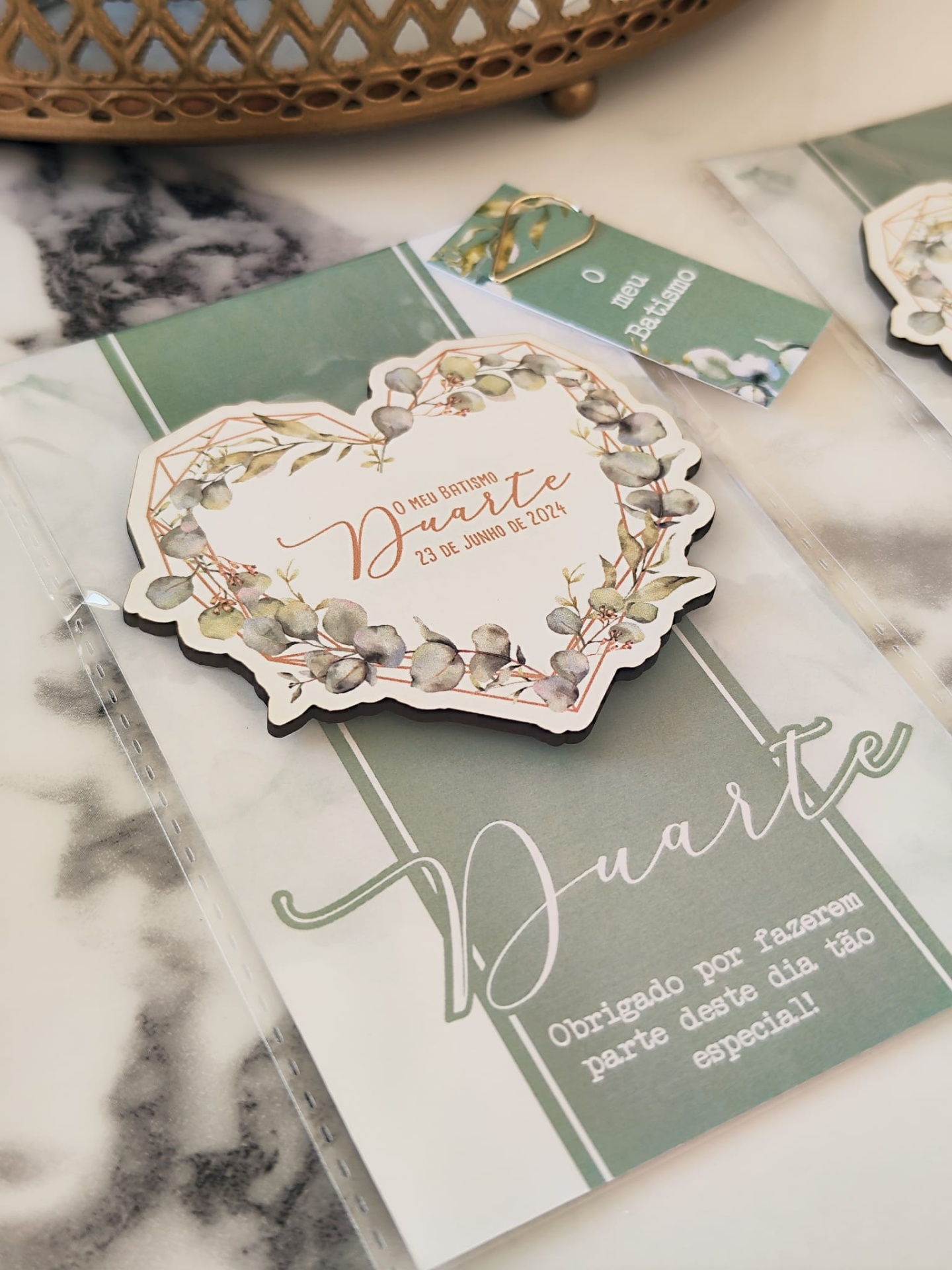 Tag de batizado em formato de coração com design floral e texto, sobre cartão verde com texto e embalagem transparente