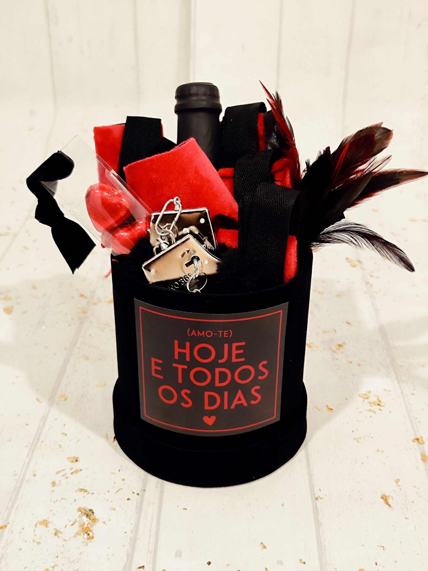 Conjunto de presentes com garrafa, penas, algemas e tecido em caixa preta com etiqueta vermelha