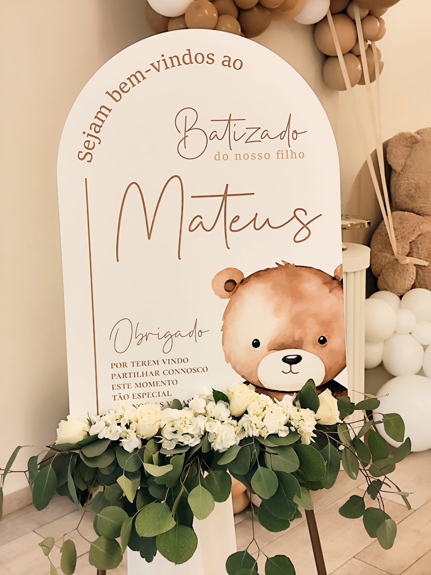 Placa decorativa para batizado com texto e desenho de urso, flores brancas e balões castanho e branco.