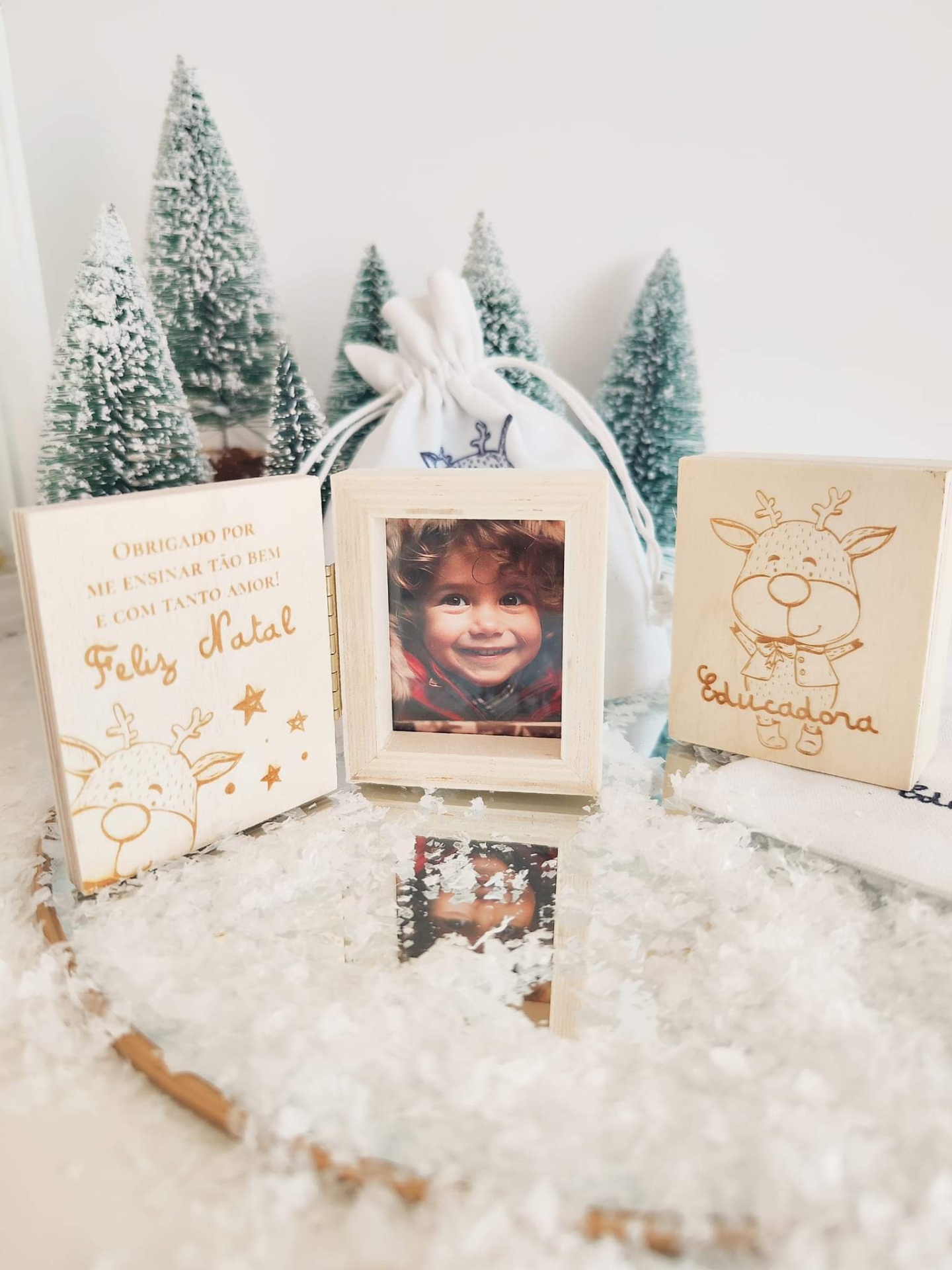 Molduras de madeira clara com desenhos de rena e texto em português, neve artificial e pinheiros nevados ao fundo