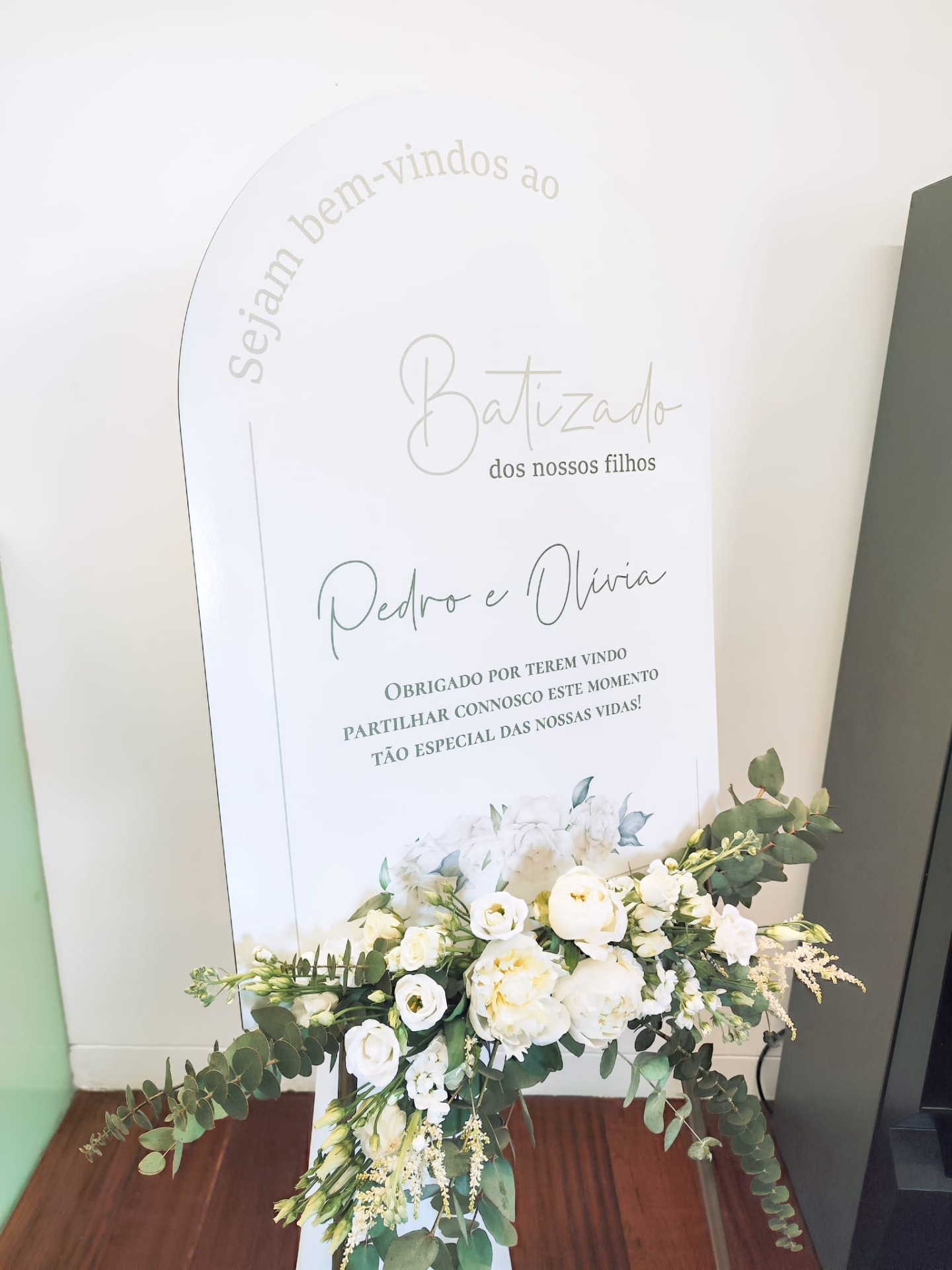 Placa branca com texto dourado e preto sobre batizado com flores brancas e verdes na base
