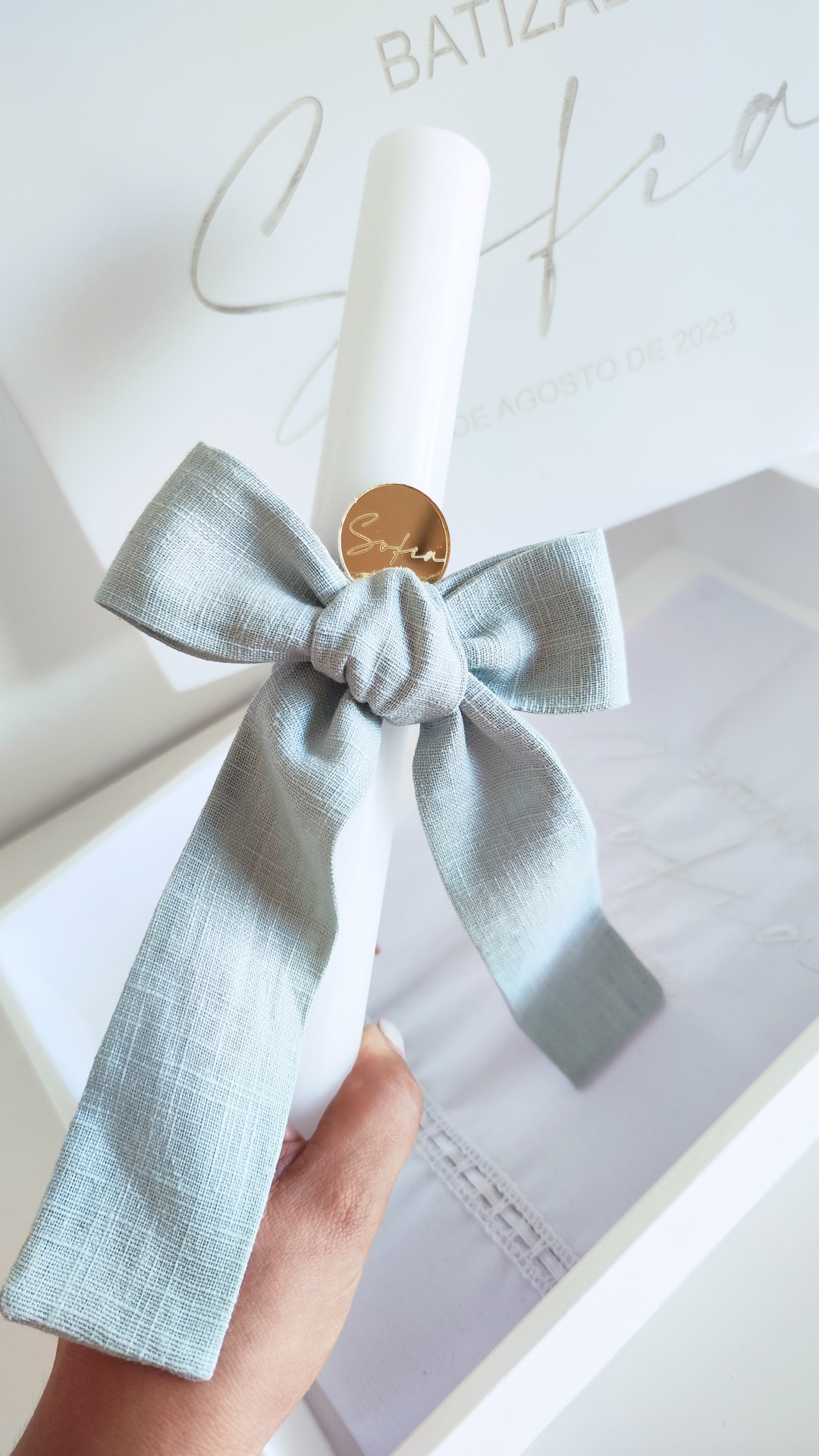 Vela branca com laço azul e etiqueta dourada em caixa com texto