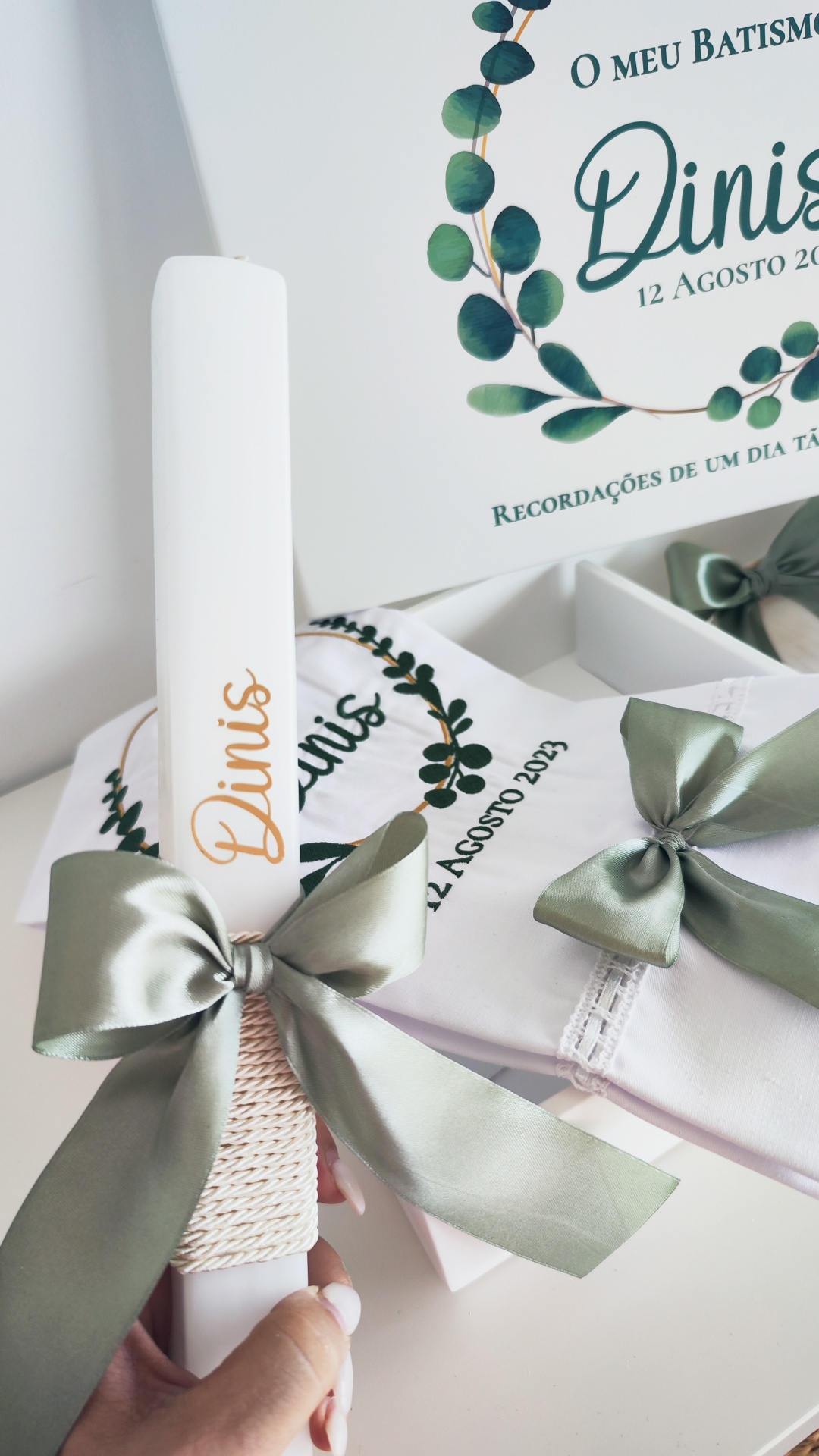 Vela decorativa branca com nome Dinis e laço verde, ao lado de pano branco bordado com texto e ilustração de folhas
