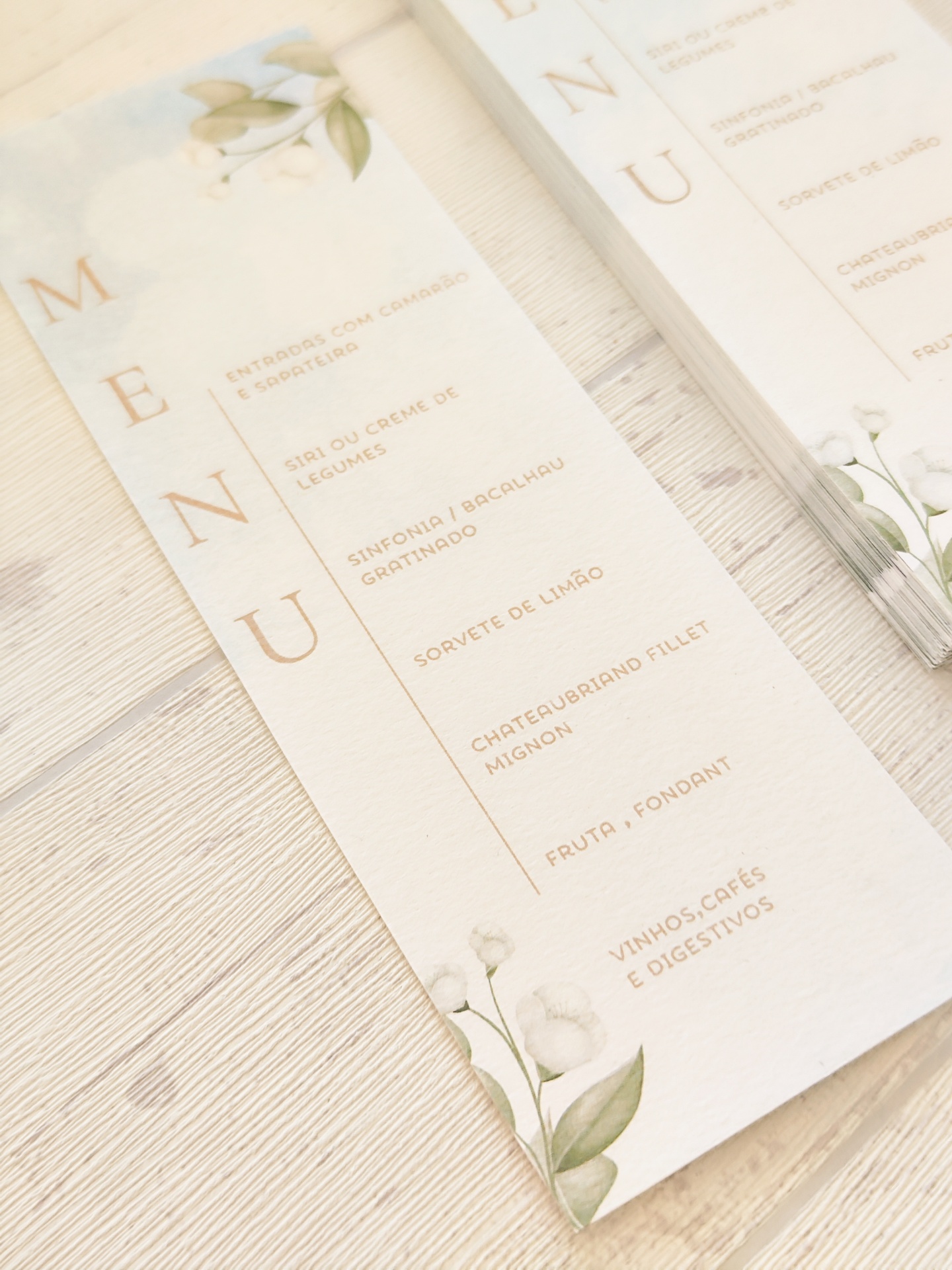 Menus em papel branco com ilustração floral sobre mesa clara.