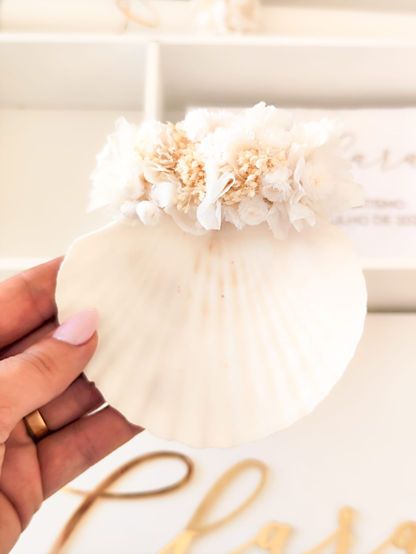Concha branca decorativa com flores secas em tons creme, segurada por mão com unhas rosa e anel dourado