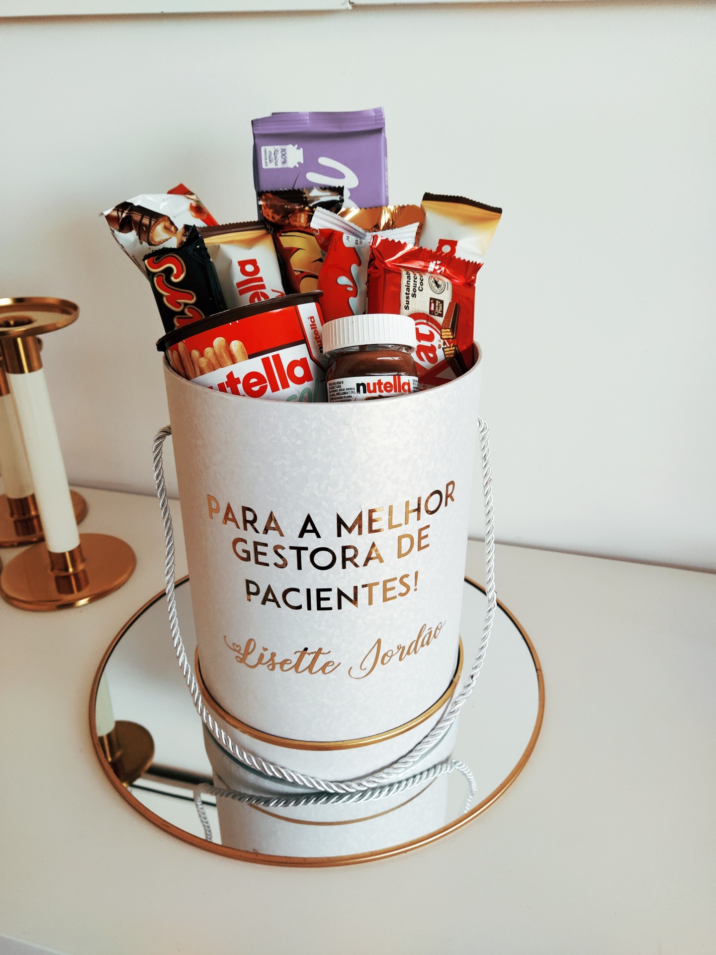 Saco branco com texto dourado e produtos Nutella e chocolates dentro, em cima de superfície branca com espelho redondo e castiçal ao lado.